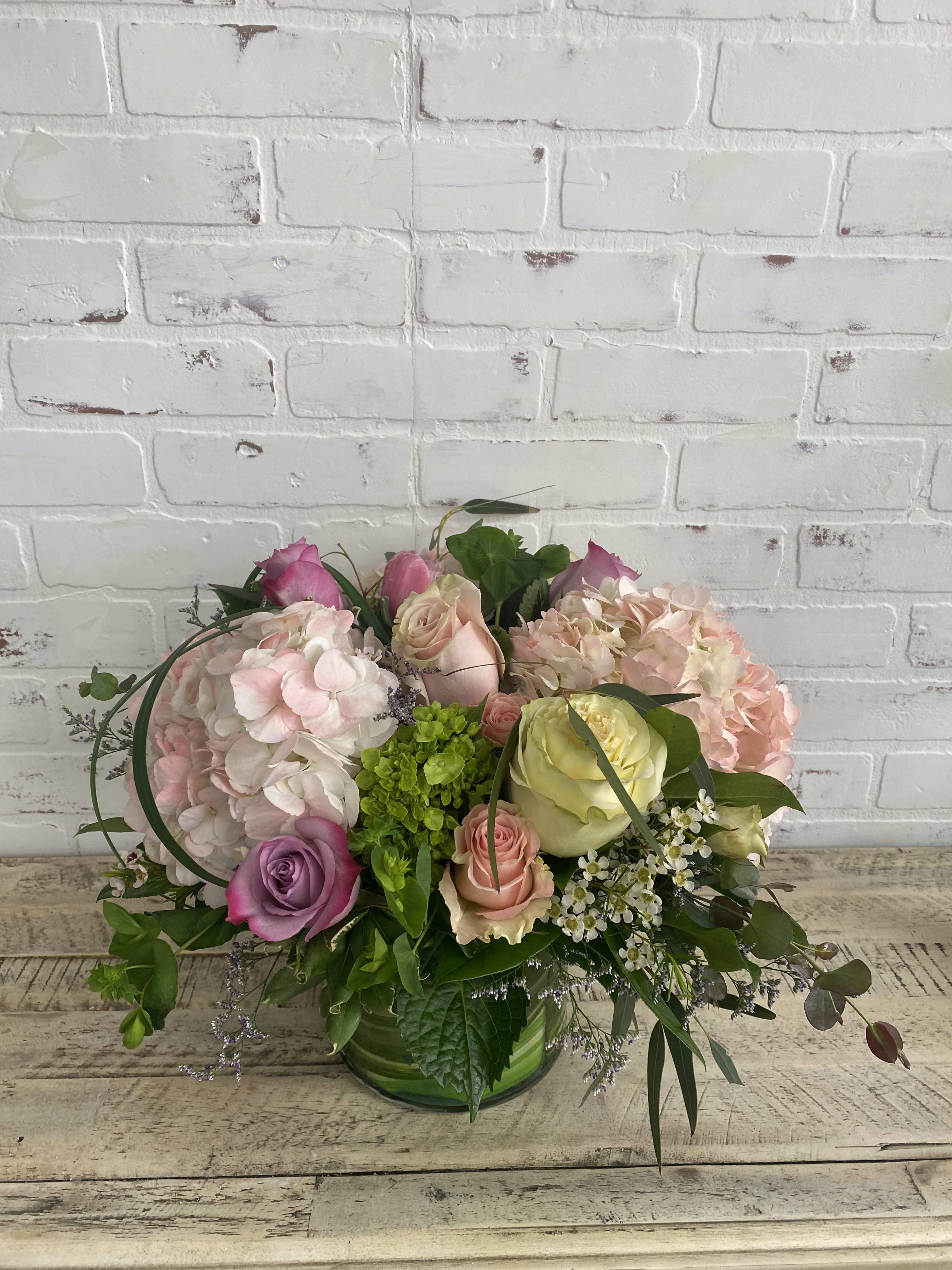 Summer wind - Light colored blooms of soft pinks, whites, green, and touches of purple. Arranged in a 6” cylinder wrapped with aspidistras leaf.  