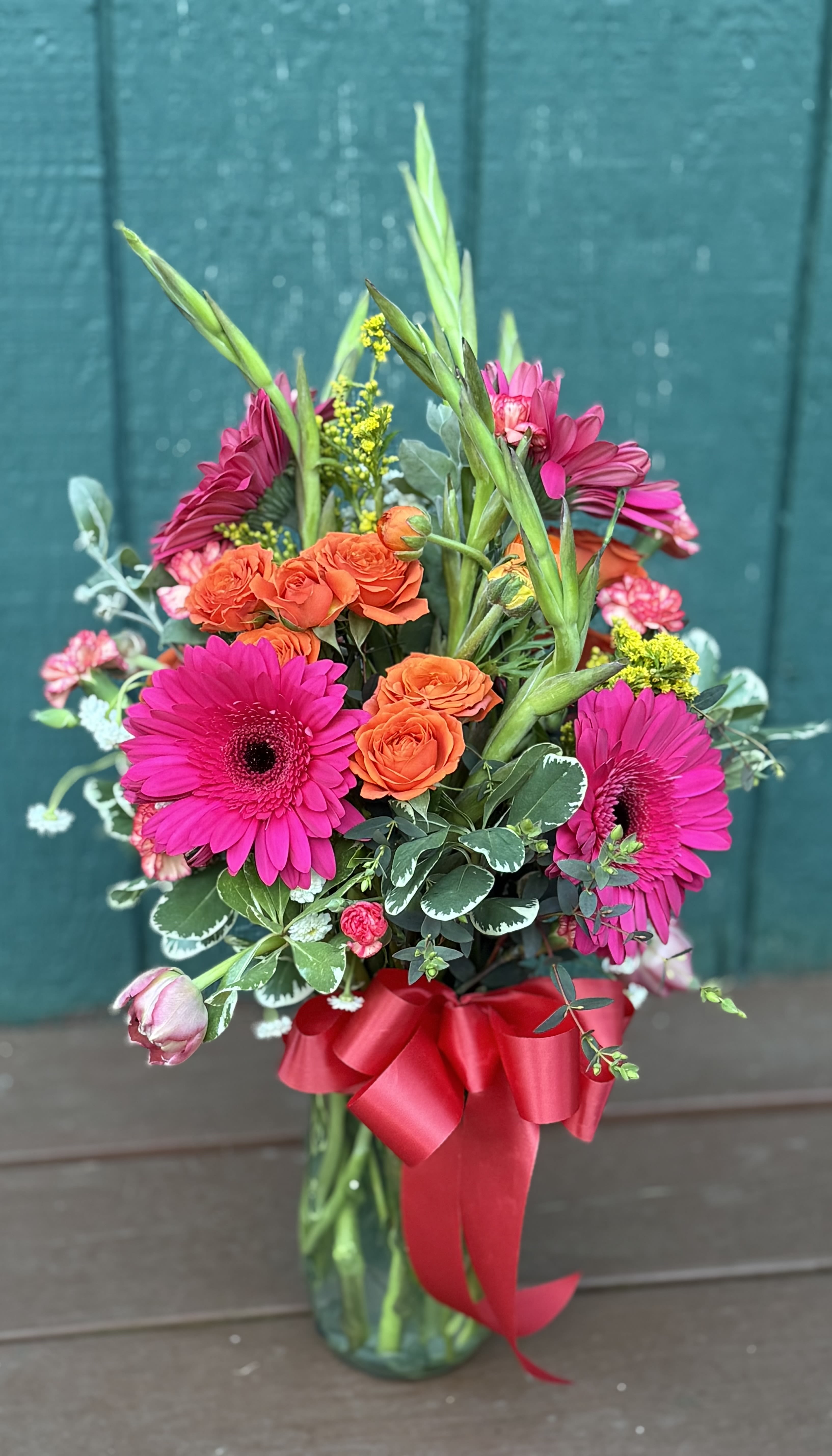 Petals &amp; Grace - This radiant arrangement is inspired by the quiet strength and joy within. Petals &amp; Grace features a bold yet graceful mix of hot pink Gerber daisies, radiant orange spray roses, delicate carnations, and soft tulips - tied with a satin ribbon and styled to celebrate both beauty and resilience. A perfect gift for someone who holds it all together with heart.   Photo is shown in Deluxe
