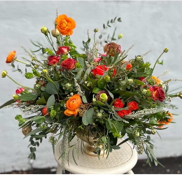 Vibrant Sunset Garden Centerpiece - Bring nature's vibrant hues to life with our Vibrant Sunset Garden Centerpiece, an exquisite arrangement designed to captivate and charm. Featuring radiant orange ranunculus, deep red spray roses, lush greenery, and delicate white accents, this floral masterpiece evokes the beauty of a glowing sunset. Perfect for celebrations, dinner parties, or simply elevating your home décor, this centerpiece is a true work of art.  Carefully designed by the skilled team at Heavenly Flowers Too, your trusted Amityville florist, this arrangement is crafted to balance bold colors with soft textures. The orange and red blooms create a warm, inviting feel, while the verdant foliage and elegant filler flowers add depth and texture. Housed in a timeless gold compote vase, this design blends classic elegance with modern sophistication.  Whether you're hosting a gathering or gifting someone special, the Vibrant Sunset Garden Centerpiece brings joy and warmth to any space. Available for flower delivery in Amityville, this one-of-a-kind arrangement ensures your sentiment arrives fresh and beautifully arranged.  Order today at Heavenly Flowers Too to experience the exceptional artistry and quality that define our floral designs. Let us help you celebrate life's moments with blooms that speak from the heart.
