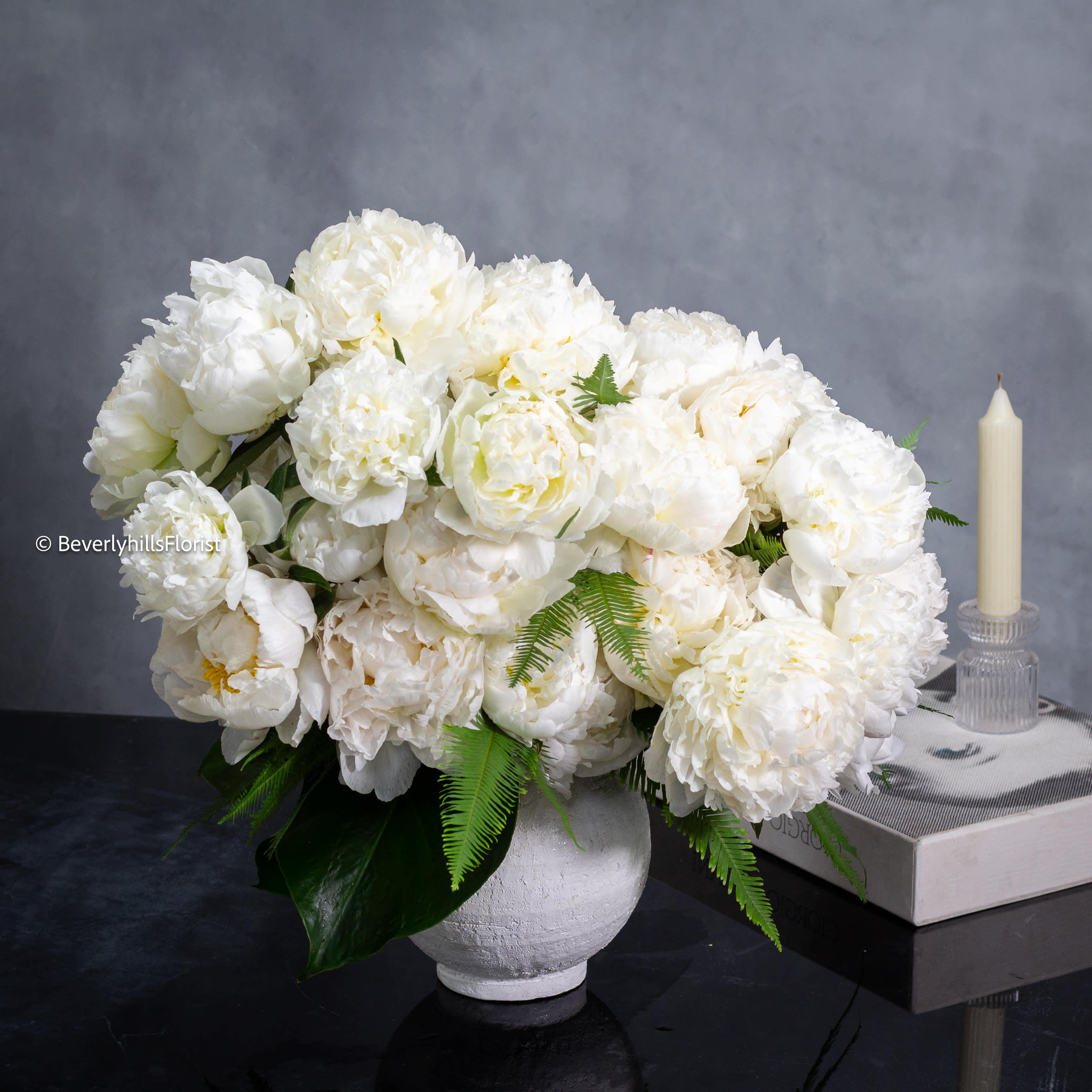 Whispering Love - A Romance in White Peonies -  A lavish arrangement of over 20 pristine white peonies nestled within a sleek 7&quot; ceramic white bowl. Each delicate bloom exudes elegance, its petals unfurling gracefully like layers of satin. The bowl serves as a pristine canvas, accentuating the purity and beauty of the blossoms it holds. Standing tall at 20 inches and spanning 23 inches wide, this arrangement commands attention with its opulent presence, a testament to nature's exquisite artistry. The lushness of the peonies, combined with the simplicity of the vessel, creates a harmonious display that is both captivating and timeless.
