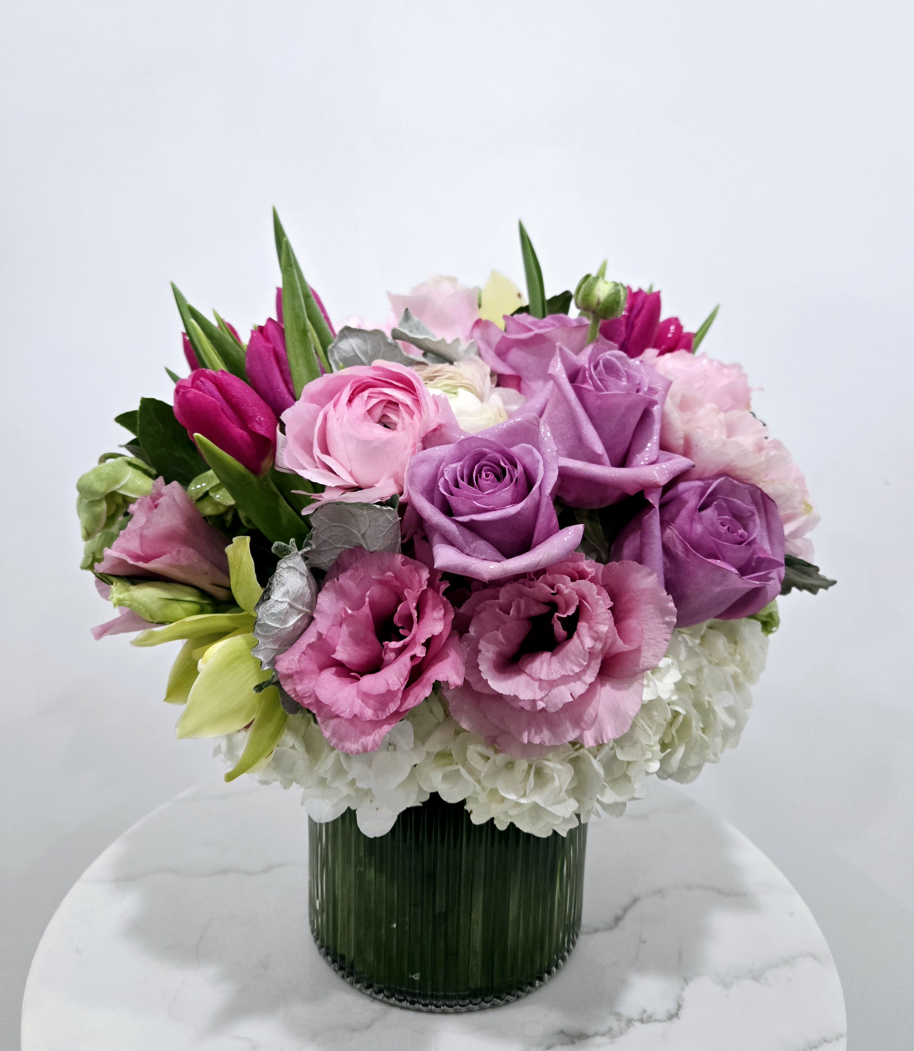 Blushing Serenity - This elegant floral arrangement, &quot;Blushing Serenity,&quot; features a soft and romantic palette of blush pinks and gentle whites. At its heart are delicate pink roses and ranunculus, symbolizing grace and new beginnings. Surrounding them are lush white hydrangeas, adding fullness and texture, while light green cymbidiums bring a refreshing pop of color. Deep pink tulips rise gracefully from the bouquet, lending a touch of boldness and movement. Set in a simple, modern vase, this arrangement evokes a sense of timeless beauty and serenity, perfect for any special occasion or serene space.