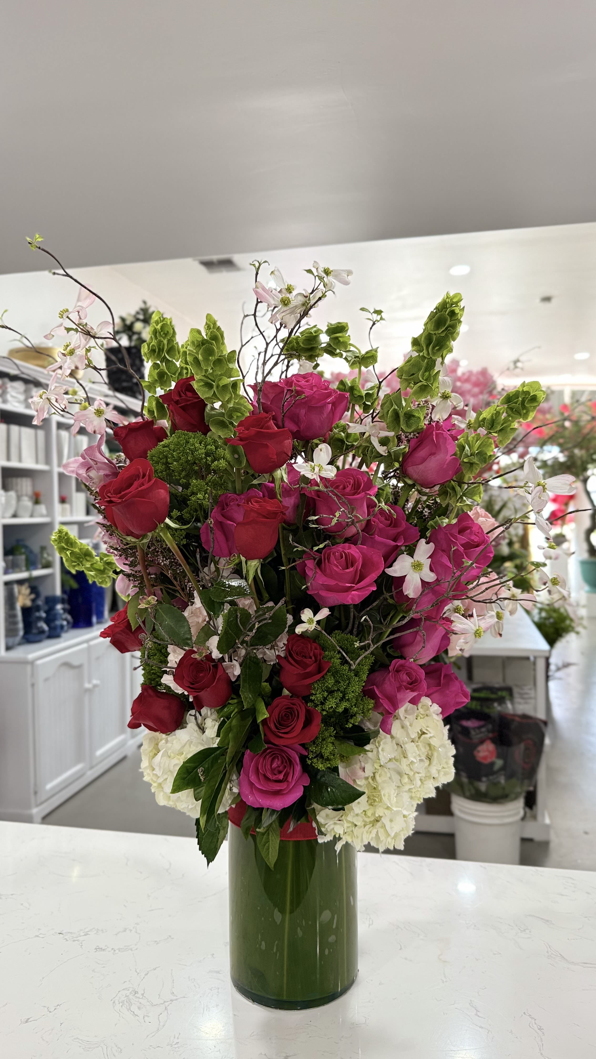 Garden Royale - Garden Royale is a grand, show-stopping arrangement bursting with rich fuchsia and red roses, delicate cherry blossom stems, lush green bells of Ireland, and soft white hydrangeas. Designed in a tall, modern vase, it brings elegance and height to any space — perfect for lobbies, events, or an unforgettable gift.