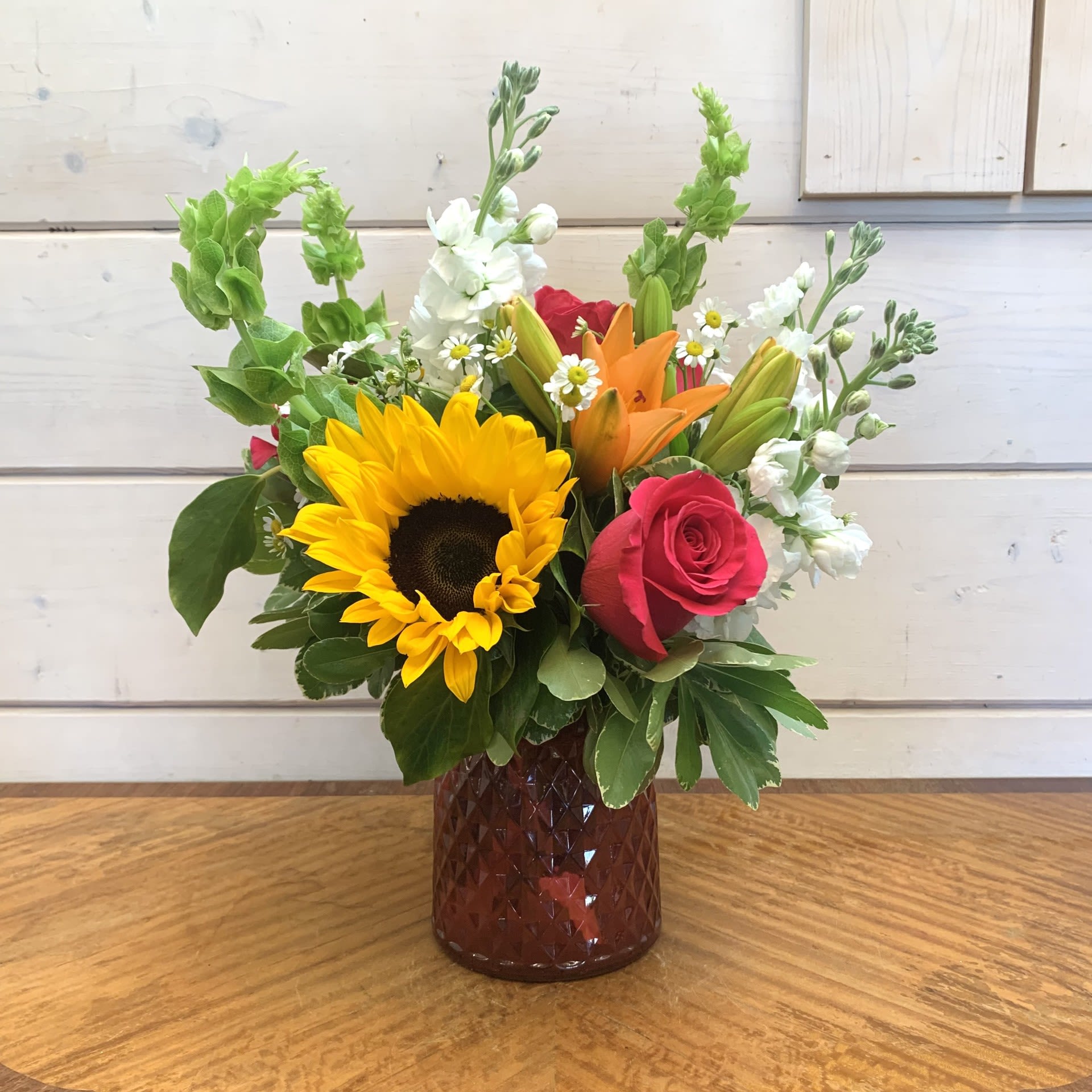 Summer Lovin' Summer Lovin' - Hot pink roses and yellow sunflowers feature with fragrant white stock, bells of ireland and orange lilies arranged in a pink cut glass posey vase with chamomile or other seasonal filler flower. Approximately 12" tall and 9" wide.