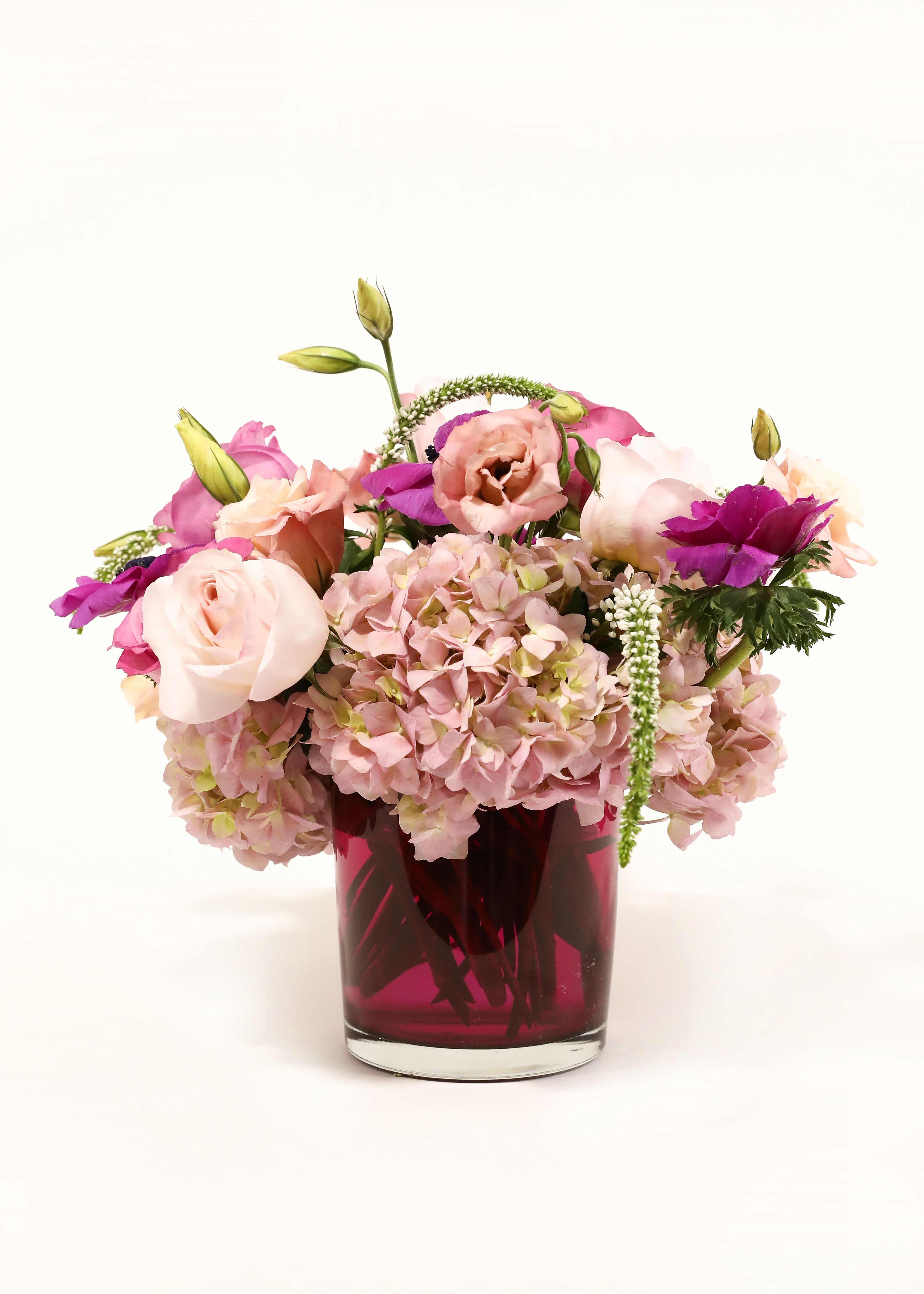 BLUSH RUSH BLUSH RUSH - THIS LOVELY PINK ARRANGEMENT HAS GARDEN ROSES, HYDRANGEA, ANENOME, LISIANTHUS, AND WHITE VERONICA IN A TRANSPARENT PINK CYLINDER GLASS VASE.