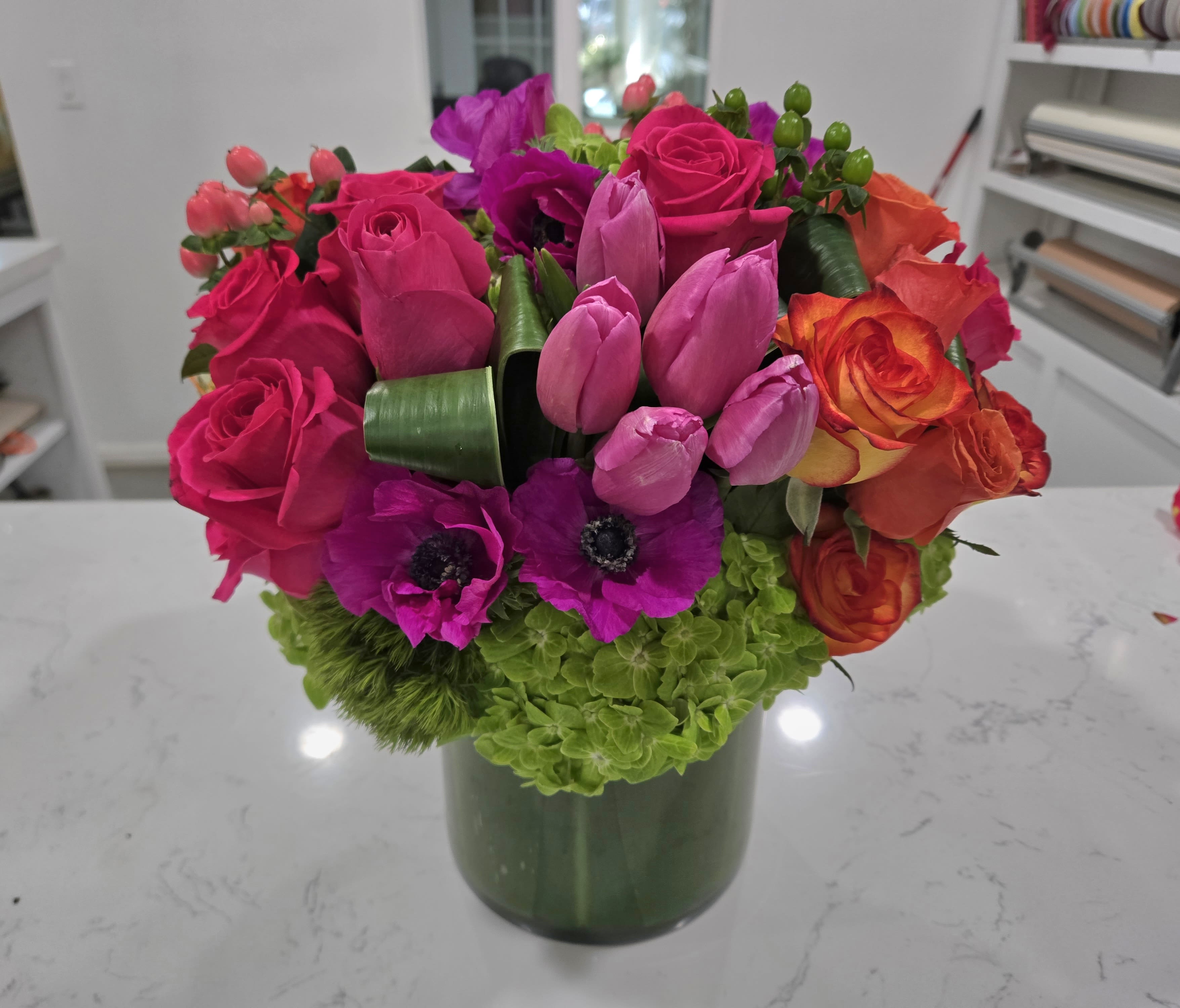Fiesta Bloom - A dazzling explosion of color, Fiesta Bloom brings vibrant energy to any space. Featuring bold magenta roses, fiery orange spray roses, deep purple anemones, and lush green hydrangeas, this striking arrangement is designed to impress. Arranged in a sleek, leaf-wrapped vase, it exudes elegance and celebration, making it the perfect statement piece for any joyous occasion.