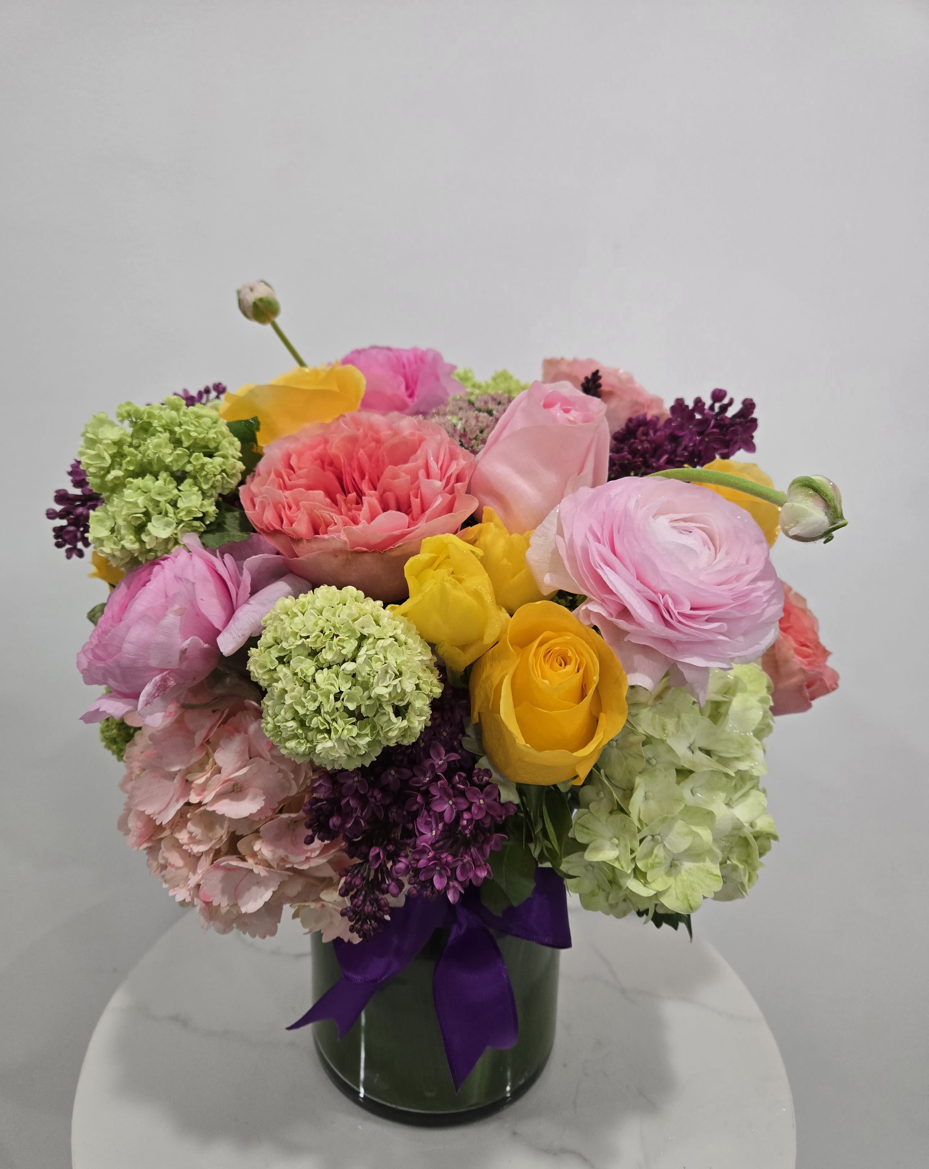 Spring Blooms - A stunning Valentine's arrangement featuring a romantic blend of roses, orchids, and seasonal blooms in warm sunset hues of pink, orange, and yellow. Accented with lush greenery and finished with a charming pink ribbon, this bouquet is a perfect expression of love and admiration for Valentine's Day.