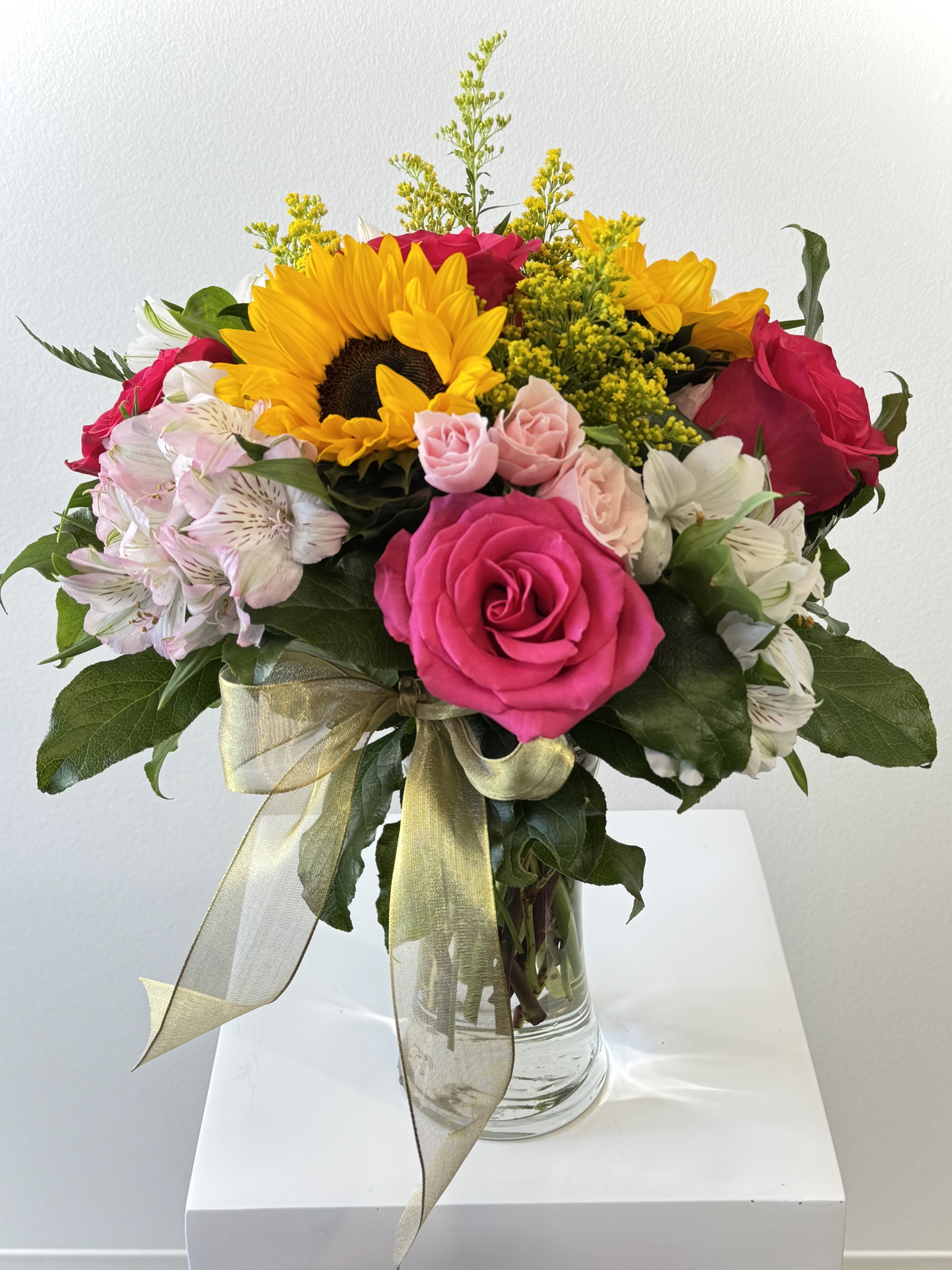 Bright and Sunny Bright and Sunny - Sunflowers, fuchsia roses, pink spray roses, solidago and alstroemeria with greenery in a glass vase finished with a gold bow