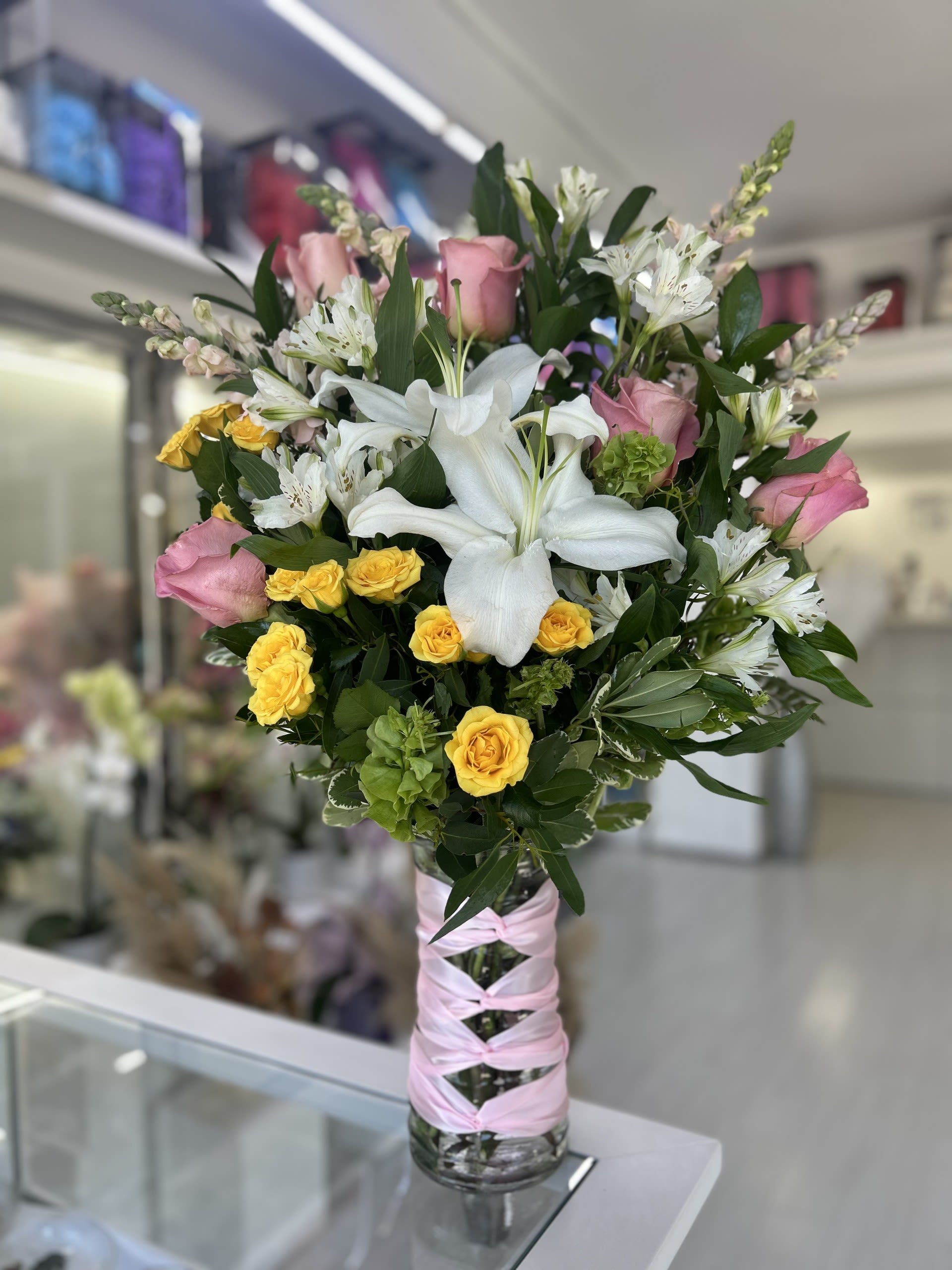 Blush Radiance - This beautiful floral arrangement features a mix of soft pink roses, vibrant yellow spray roses, with elegant white lilies and alstroemerias, all tied together with a pink ribbon!  If some flowers and/or the vase are not available there will be minor substitutions