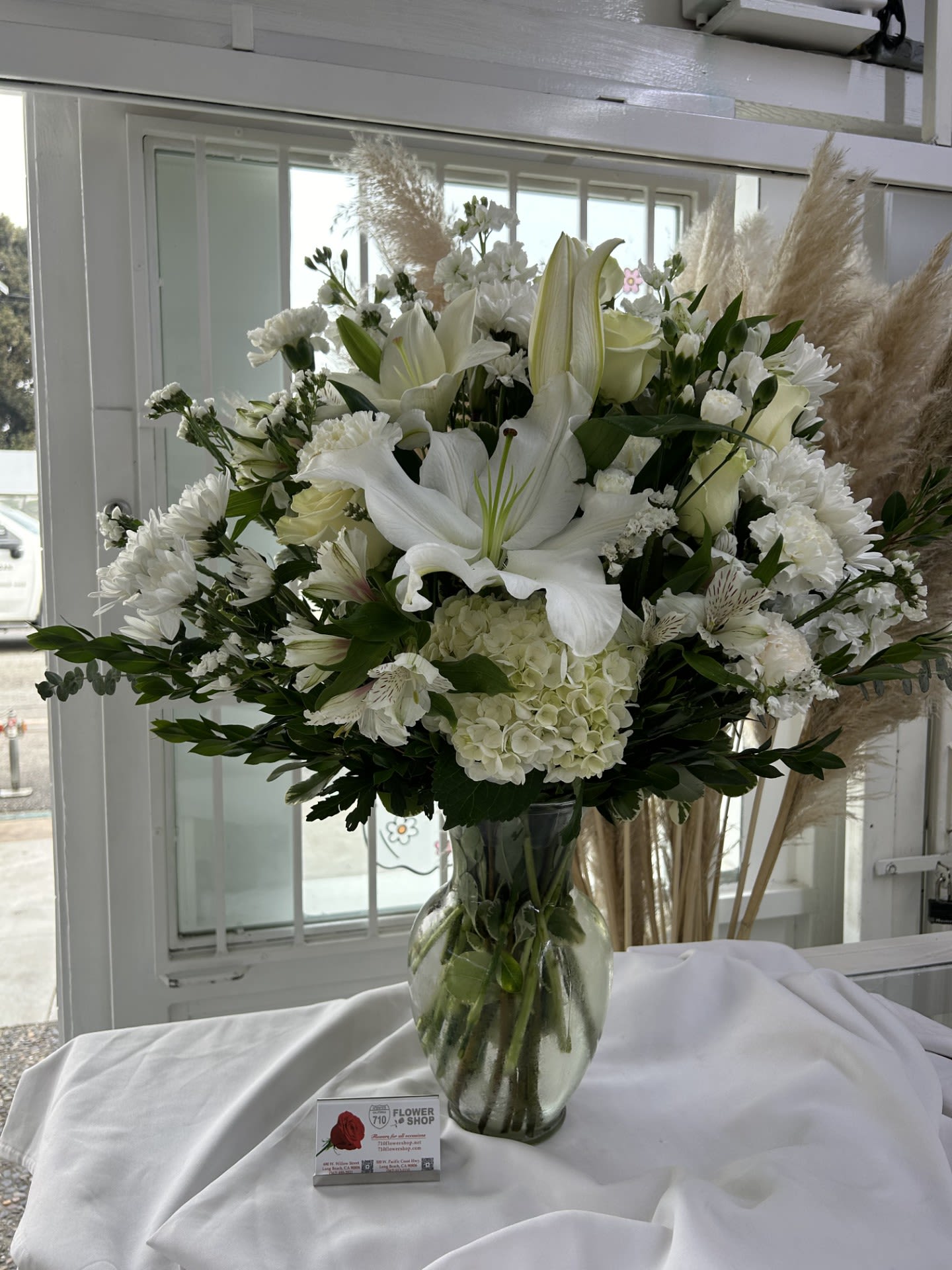 Pure Serenity - The delicate white lilies, roses, and soft greenery create a calming and peaceful atmosphere that radiates simplicity and grace. If some flowers and/or the vase are not available there will be minor substitutions.