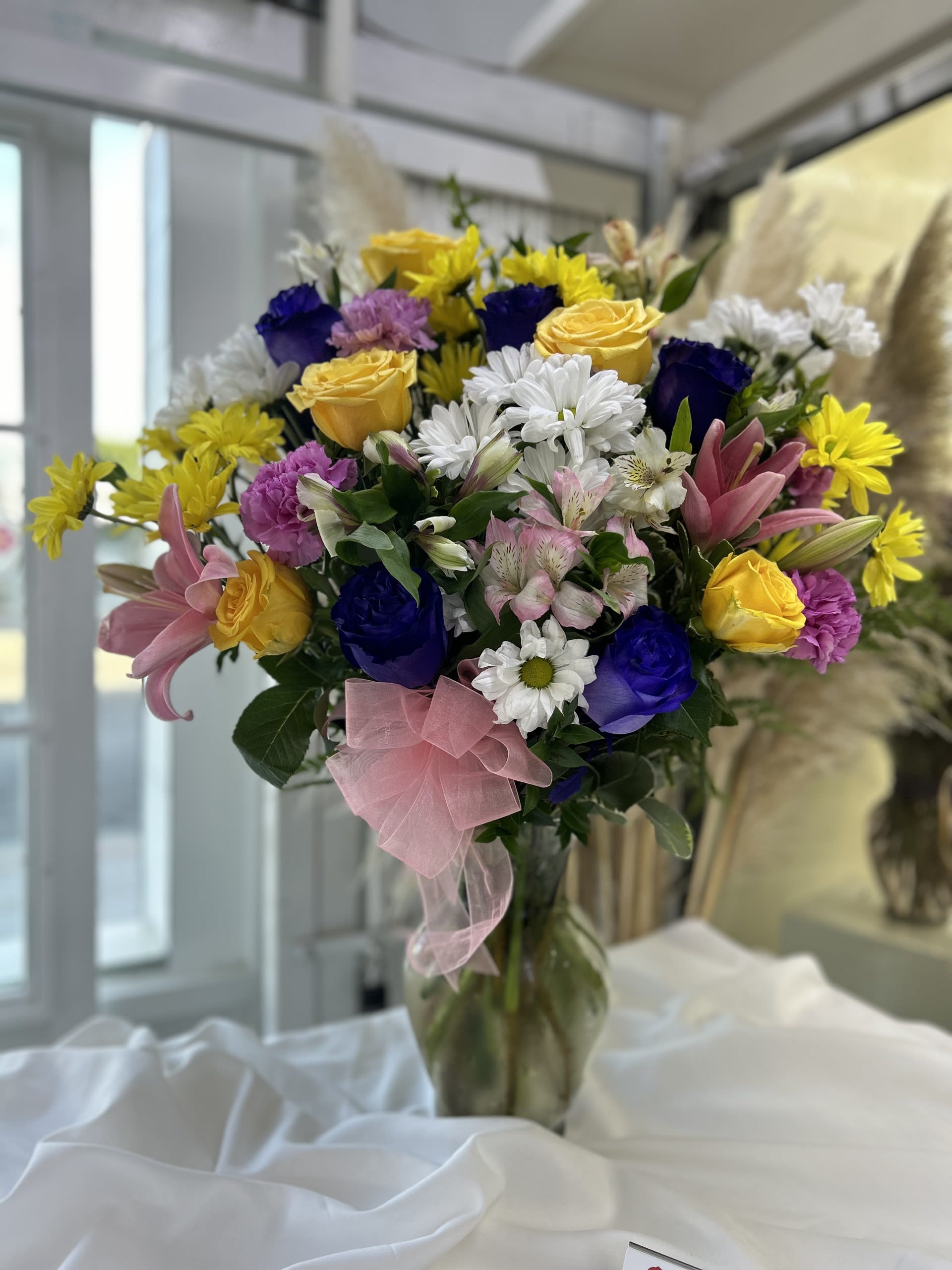 Joyful Radiance - The mix of vibrant blues, yellows, pinks, and purples, along with the soft pink bow, gives off a cheerful and lively energy that radiates happiness. Substitutions: If some flowers and/or the vase are not available there will be minor substitutions.  
