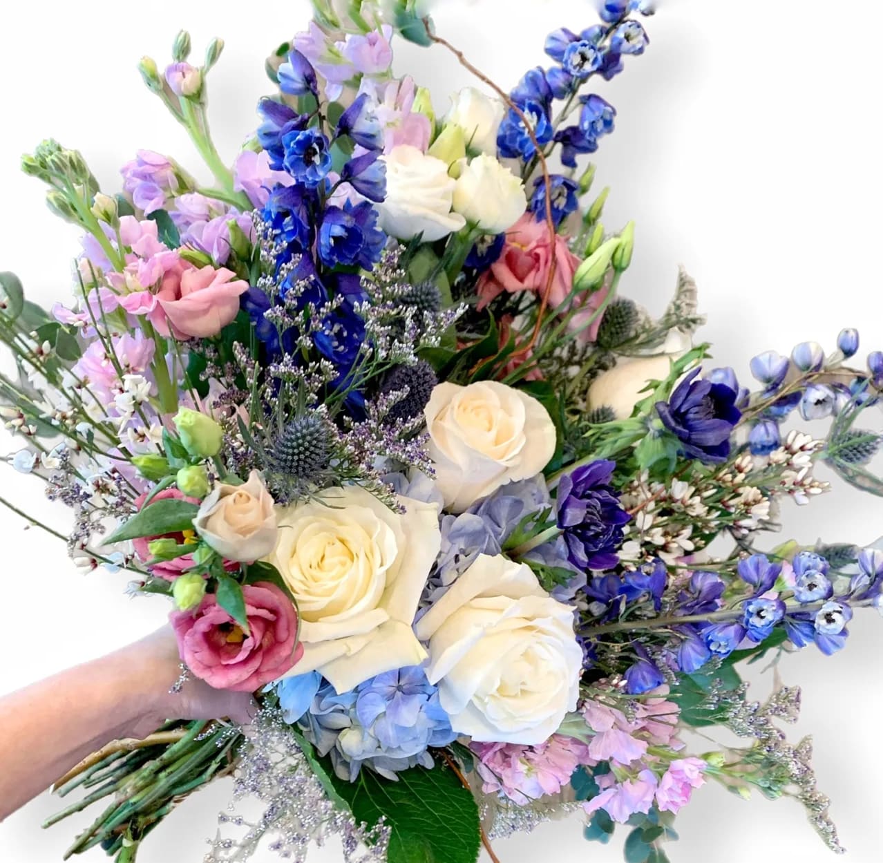 Hand Wrapped - Blues, White, Pink, Lavender - A hand wrapped bouquet of gorgeous deep blue delphiniums with white premium long stem roses, accented with lisianthus and seasonal greenery. The bouquet comes wrapped in our signature packaging with a ribbon.  *Pictured is STANDARD.