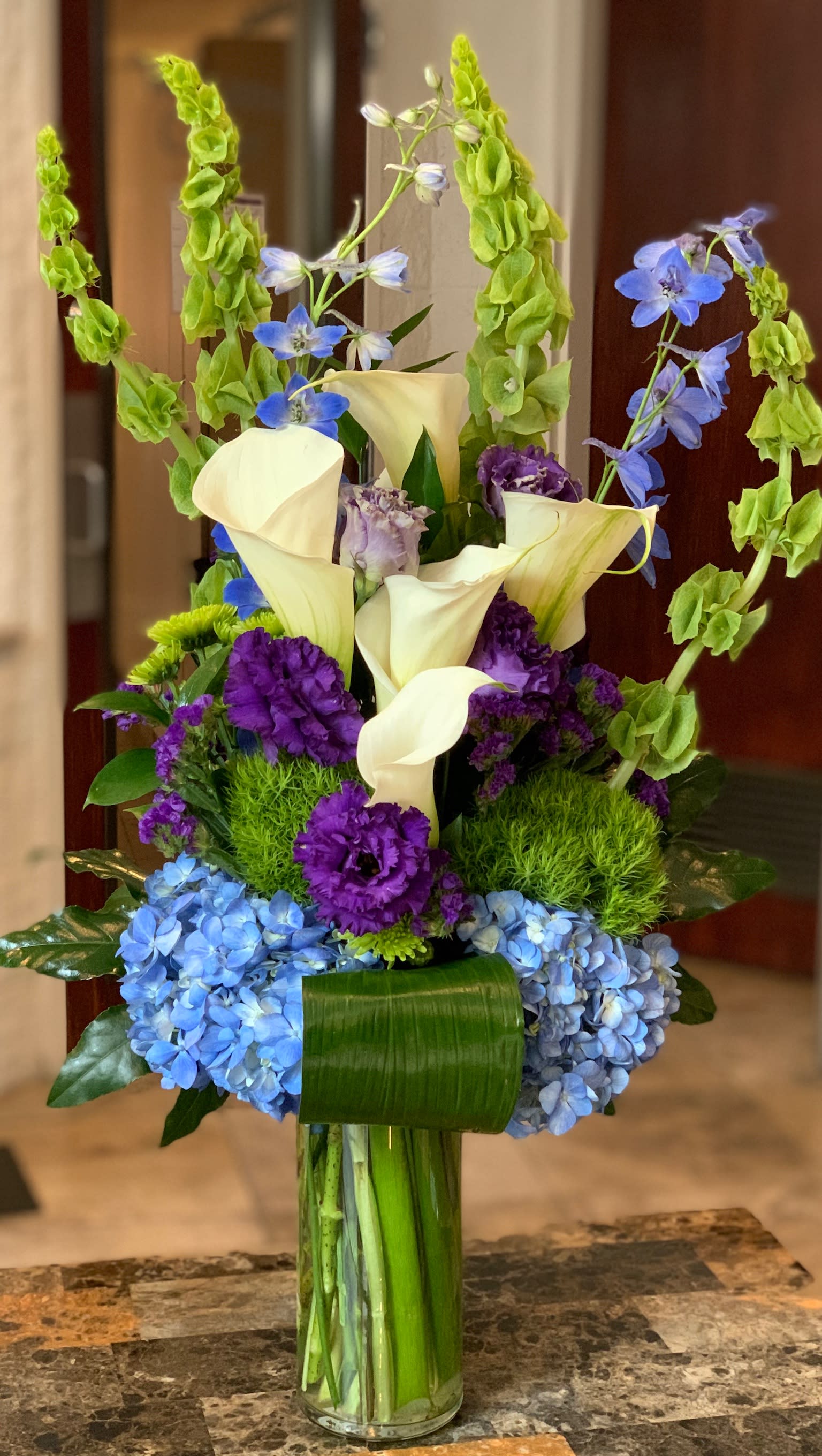Lavender and Blue Mixed Flower Bouquet  - White calla lilies, blue hydnrageas, green molucella, purple lisianthus, green dianthus, blue belladonna, purple carnations, green tropical leaves arrives in a clear glass vase.