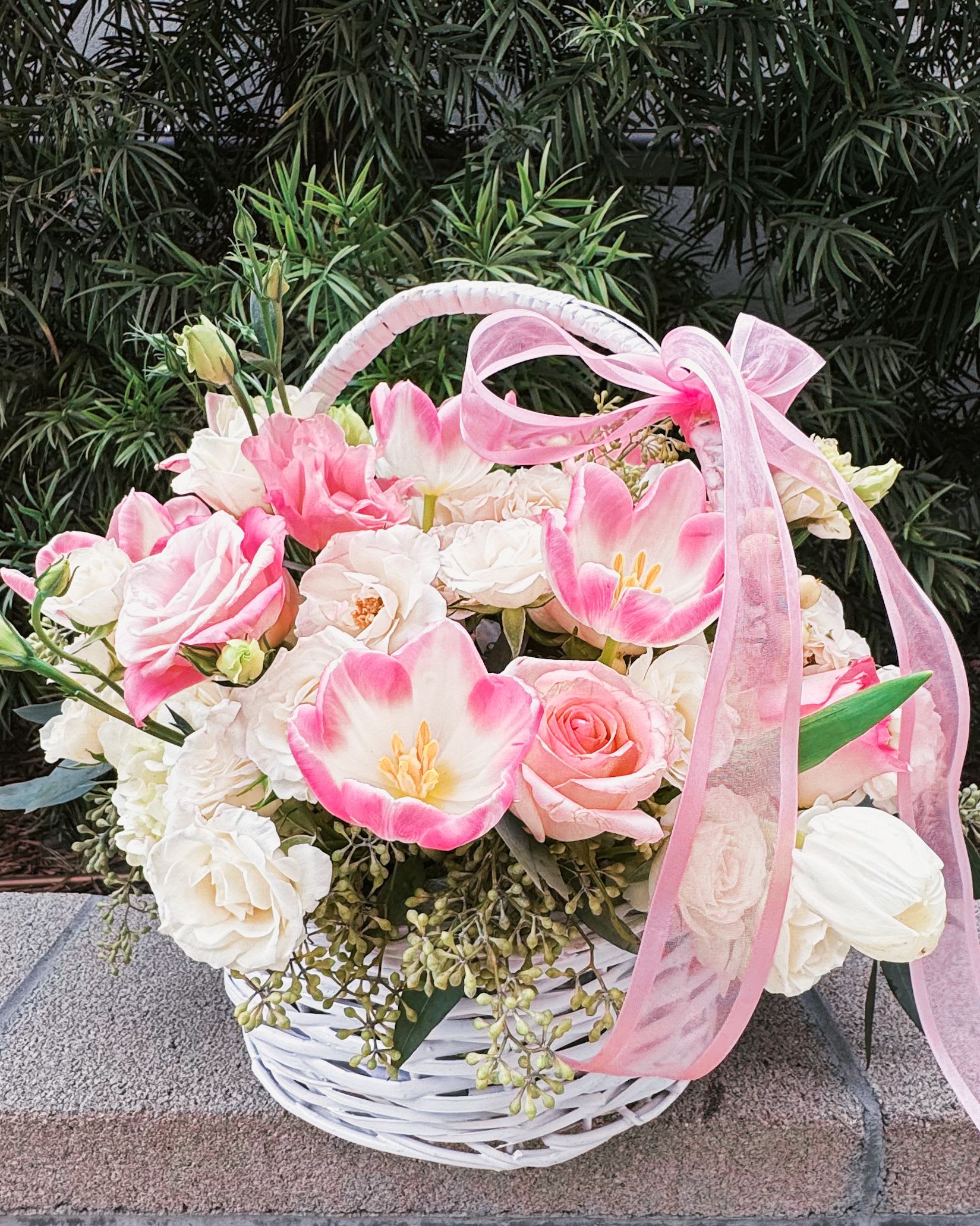 Shades of Caress - This charming basket bouquet brims with delicate pink roses, creamy white spray roses, and cheerful pink tulips, creating a soft, romantic harmony. Accented with lush white hydrangeas, lisianthus buds, eucalyptus leaves, and finished with a dainty pink ribbon, it is a perfect symbol of elegance and grace.  Flowers and Décor: Pink roses: Representing love and sweetness. White spray roses: Adding charm and delicacy. Pink tulips: A cheerful and tender touch. White hydrangeas: Offering volume and softness. Lisianthus buds: Adding subtle sophistication. Eucalyptus leaves: Bringing freshness and greenery. White wicker basket and pink ribbon: Completing the design with rustic elegance.