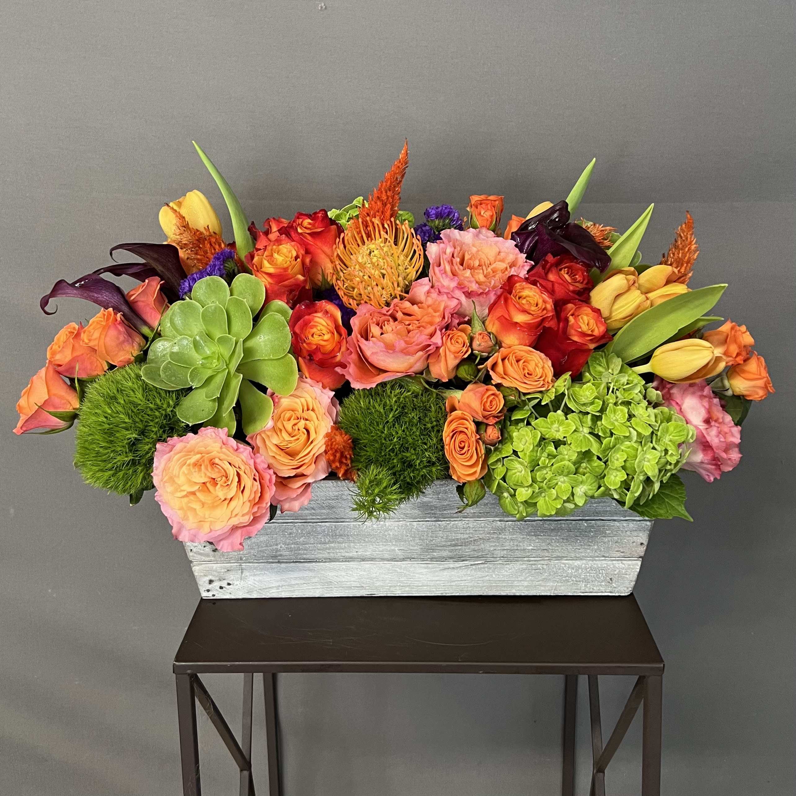 ORANGE BLOOM - This flower arrangement is designed in a wood vase and has a mix of roses and seasonal flowers. the wood vase measures 14”L x 5” W x 4” D. 