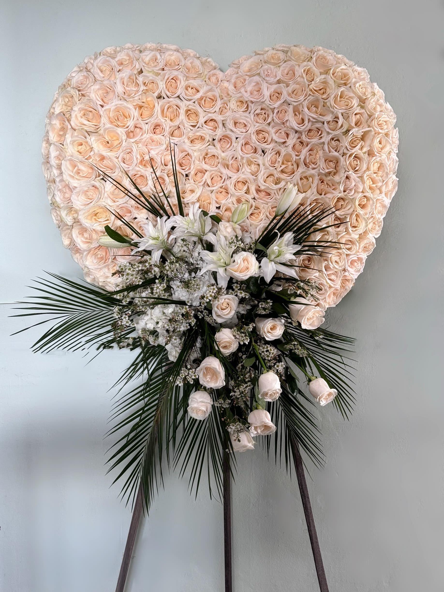 Everlasting Devotion Heart Everlasting Devotion Heart - A stunning blush rose heart tribute, delicately designed with soft peach and ivory roses, accented by white lilies and cascading greenery. This standing arrangement expresses deep love, admiration, and sympathy, making it a meaningful tribute for memorial services.