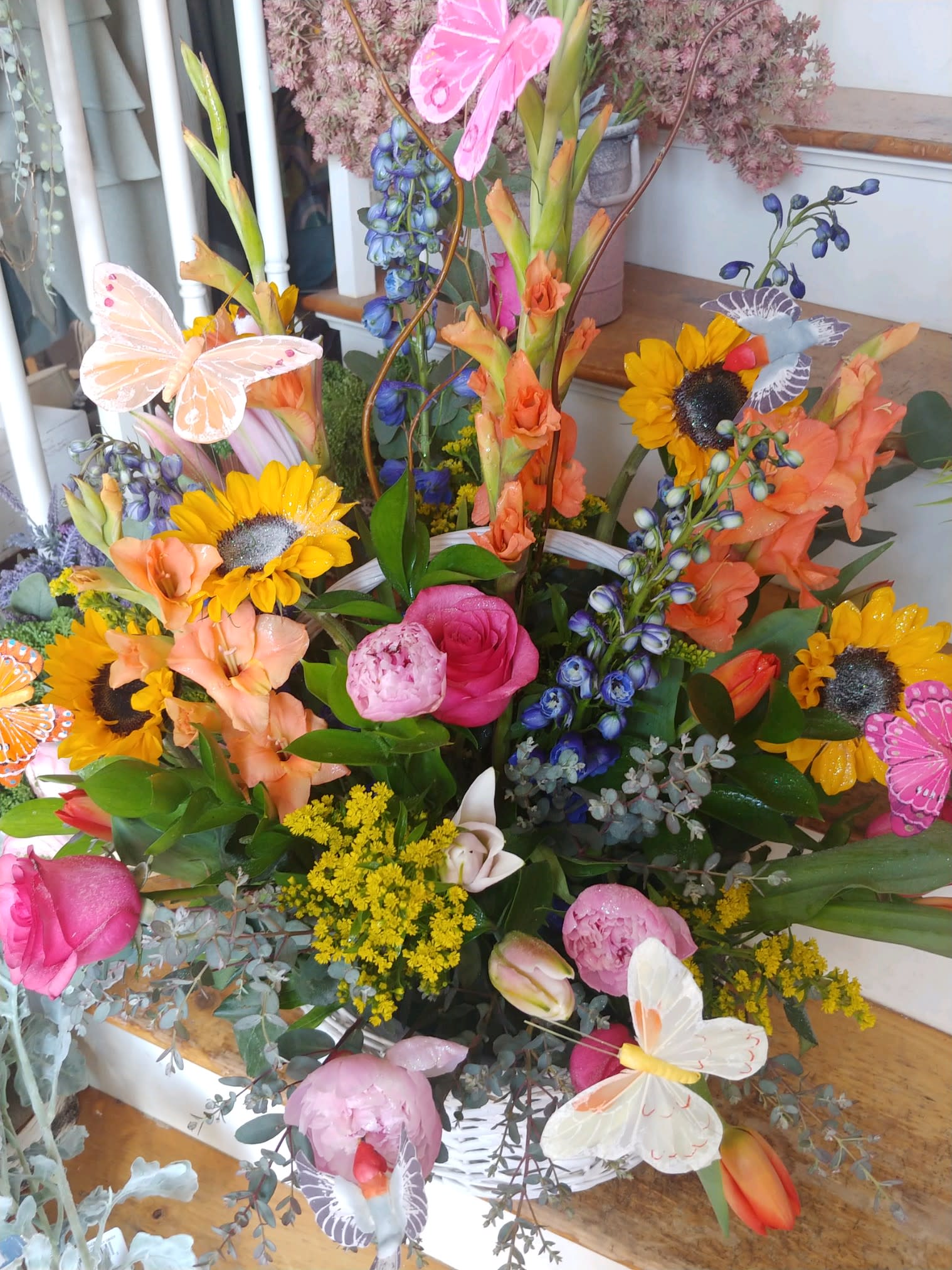 Whimsical Butterfly Garden Basket Whimsical Butterfly Garden Basket - Blue delphinium, sunflowers, peonies, roses, tulips, glads, lilies, solidago, butterflies and assorted greens arranged in a wicker basket