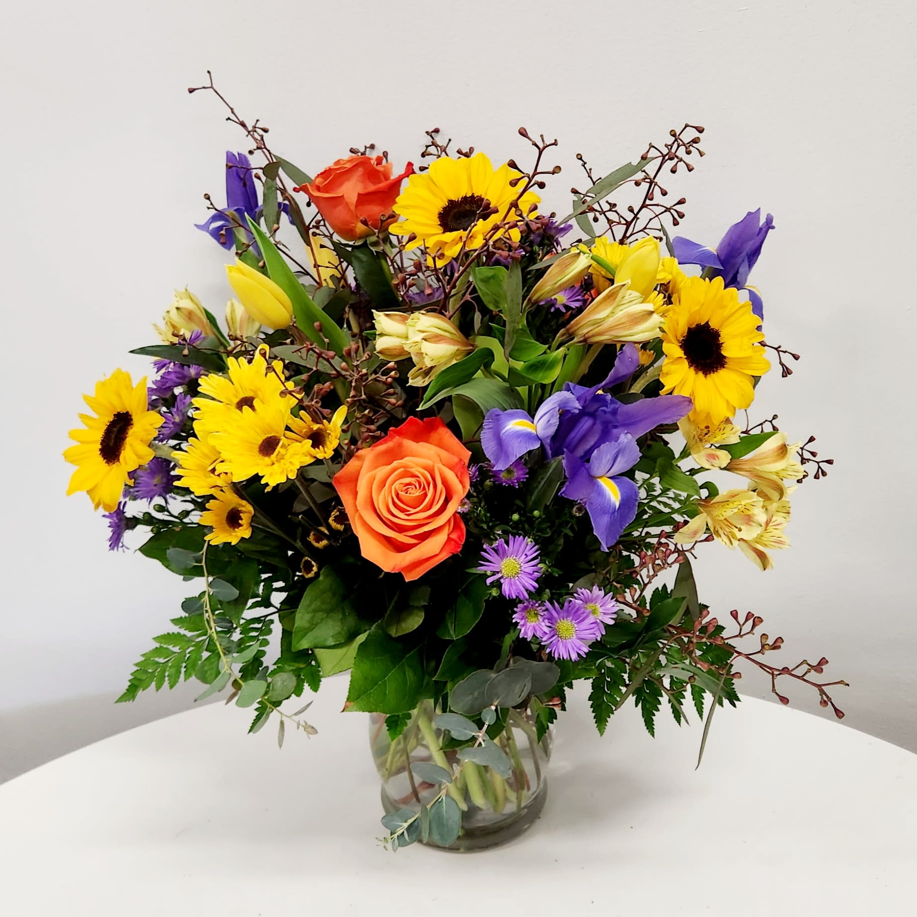 KMD2503 Free Spirit Mom KMD2503 Free Spirit Mom - KMD2503 Free Spirit Mom Bright, joyful, and full of personality—just like her. Free Spirit Mom is a cheerful mix of blooms that feels like a fresh walk through a sunny spring meadow. At the heart of this vibrant arrangement is the beloved Free Spirit Rose, with its enchanting blend of peach and orange tones. Surrounding it are deep blue irises, sunny yellow daisies, bold sunflowers, tulips, green mini hydrangeas, and other seasonal favorites. It’s a burst of happiness in every stem, designed for the mom who brings light, laughter, and a bit of whimsy wherever she goes. Celebrate her colorful spirit with flowers that match her energy.