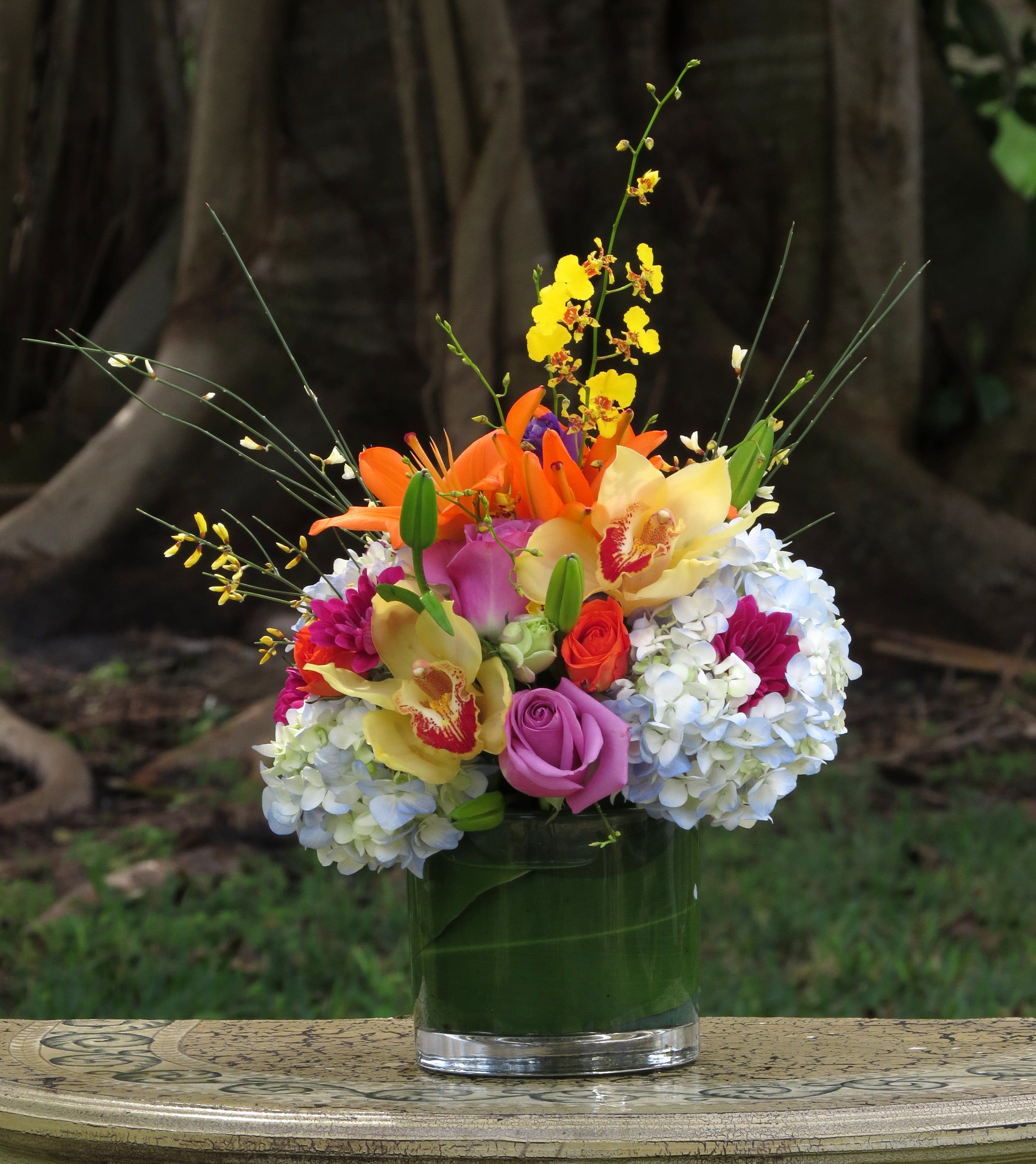 Blossoms In Vogue - High-fashion flowers for the style-minded! This modern presentation of multicolored orchids, roses, lilies and blue hydrangea has a tropical feel thanks to the bright color palette and the aspidistra leaves in the vase.