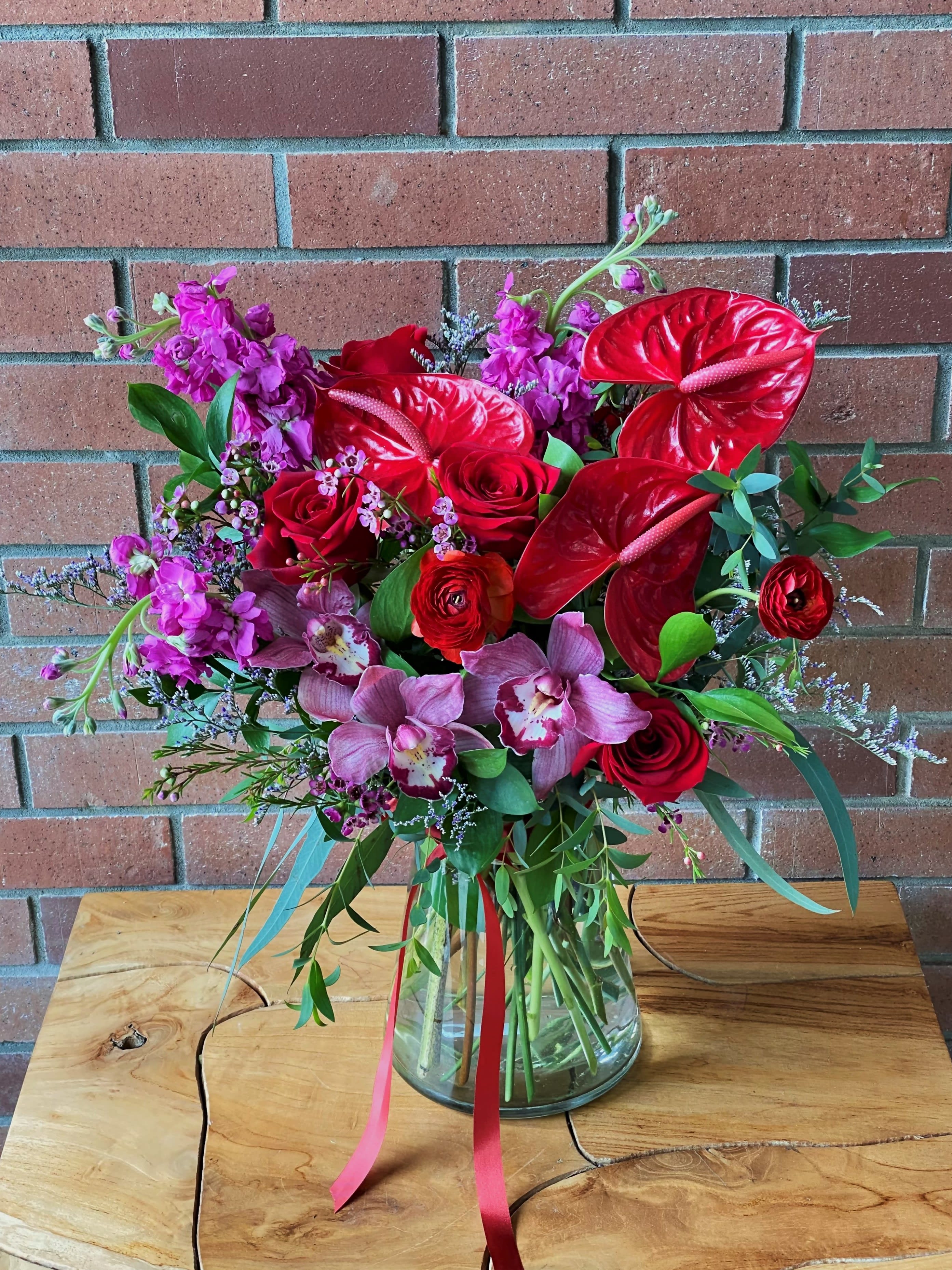 Ruby Radiance Bouquet -  Elegant reds and pinks to create this wonderful display for that special loved one. Red roses, cymbidium orchids, anthurium, fragrant stock, ranunculus arranged with premium fillers and greens in a crystal clear vase.