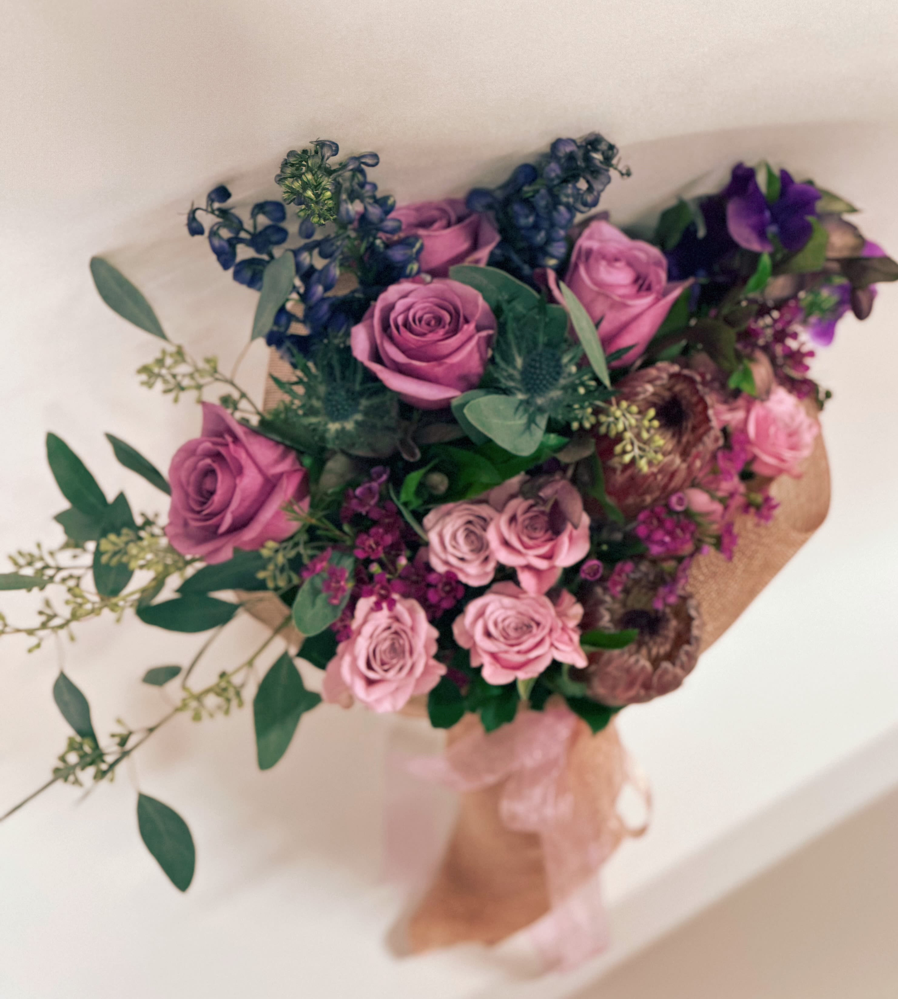 Purple Hues Wrapped Fresh Bouquet - This seasonal fresh bouquet by Venetian flowers is a stunning arrangement that beautifully showcases a variety of purple hues along with soft pink accents. At the heart of the bouquet are rich purple roses, complemented by fragrant lavender and vibrant purple stock, which add depth and texture. The striking purple delphinium reaches upward, introducing a touch of elegance and height to the composition.  Nestled among these blooms are delicate pink ice proteas, which bring a unique and exotic flair, while soft spray roses add a charming, romantic feel. Eucalyptus leaves lend a refreshing greenery, enhancing the bouquet's overall fragrance and visual appeal. Sweet peas, with their sweet scent and dainty blossoms, weave through the arrangement, adding a whimsical touch.  The entire bouquet is artfully wrapped in an organic mesh flower wrap, allowing the beauty of the flowers to shine through while providing a natural, rustic look. Sheer ribbons in complementary colors cascade gracefully from the wrap, completing this delightful and seasonal floral masterpiece.