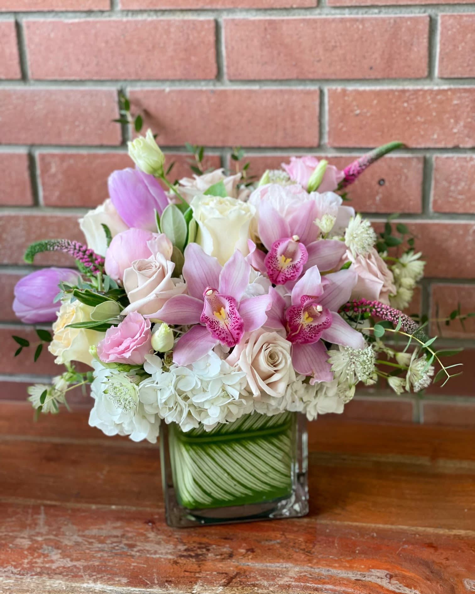 Blush Garden Blush Garden - Pink Mini Cymbidiums, White Hydrangeas, Blush Colored Roses, Tulips, Veronicas, Lisianthus in a modern 4x4 square cube wrapped with variegated tropical leaves. 12” bouquet size.