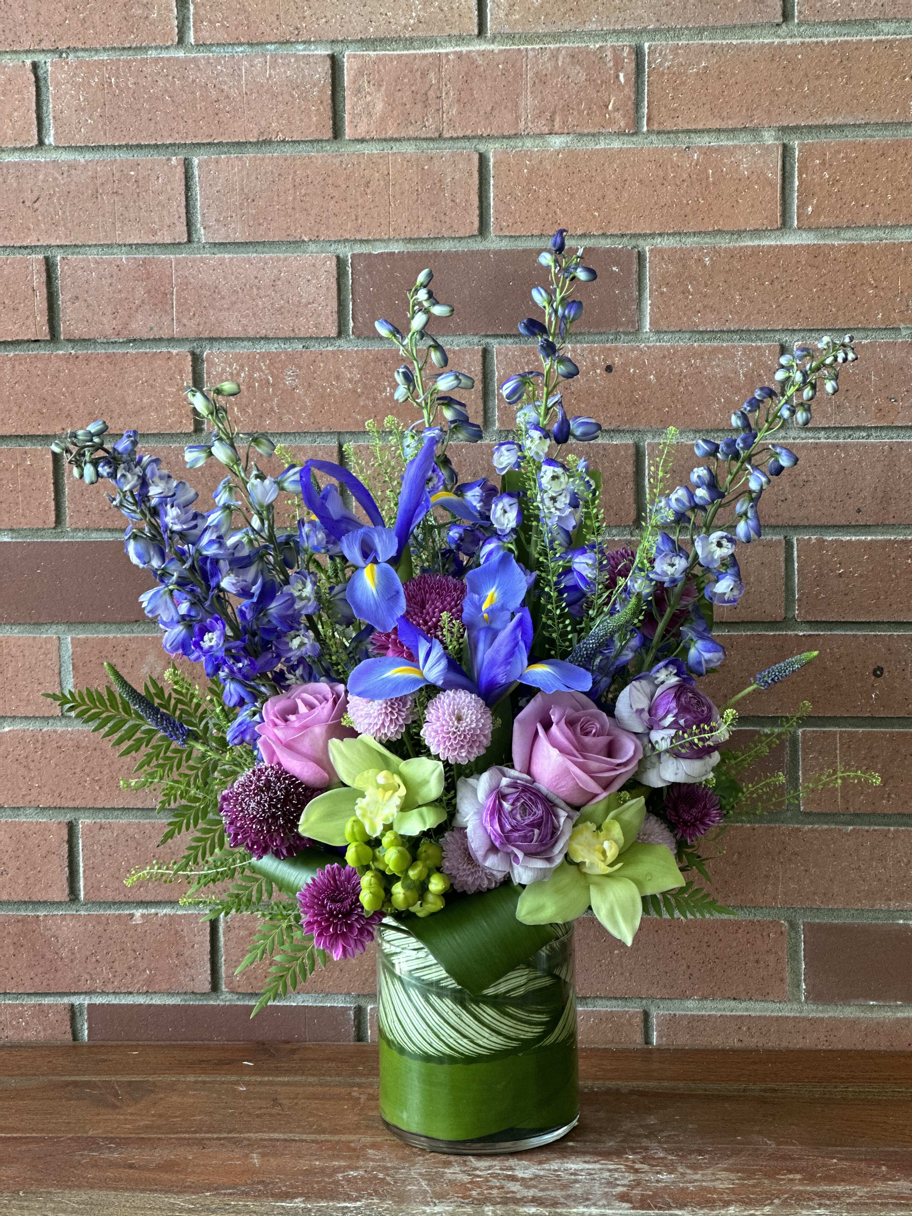 Shades Of Purple &amp; Lime Bouquet  - This lovely bouquet filled with purple delphinium, blue iris, purple mums, lavender roses, beautiful purple ranunculus and lime green orchids, hypericum berries and lush lime greenery accented with tropical leaves designed in a beautiful modern cylinder vase. 