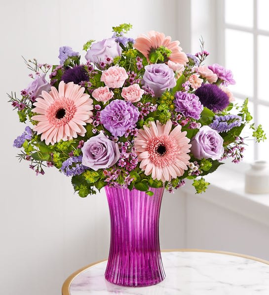 Sweet Affection - The arrangement with lavender roses, carnations and statice; pink Gerbera daisies and waxflower; light pink mini carnations; purple trachelium; accented with assorted greenery Artistically designed in our exclusive fluted gathering vase in a lovely shade of violet; vintage pressed glass with ribbed detail creates a charming enhancement for the floral arrangement;