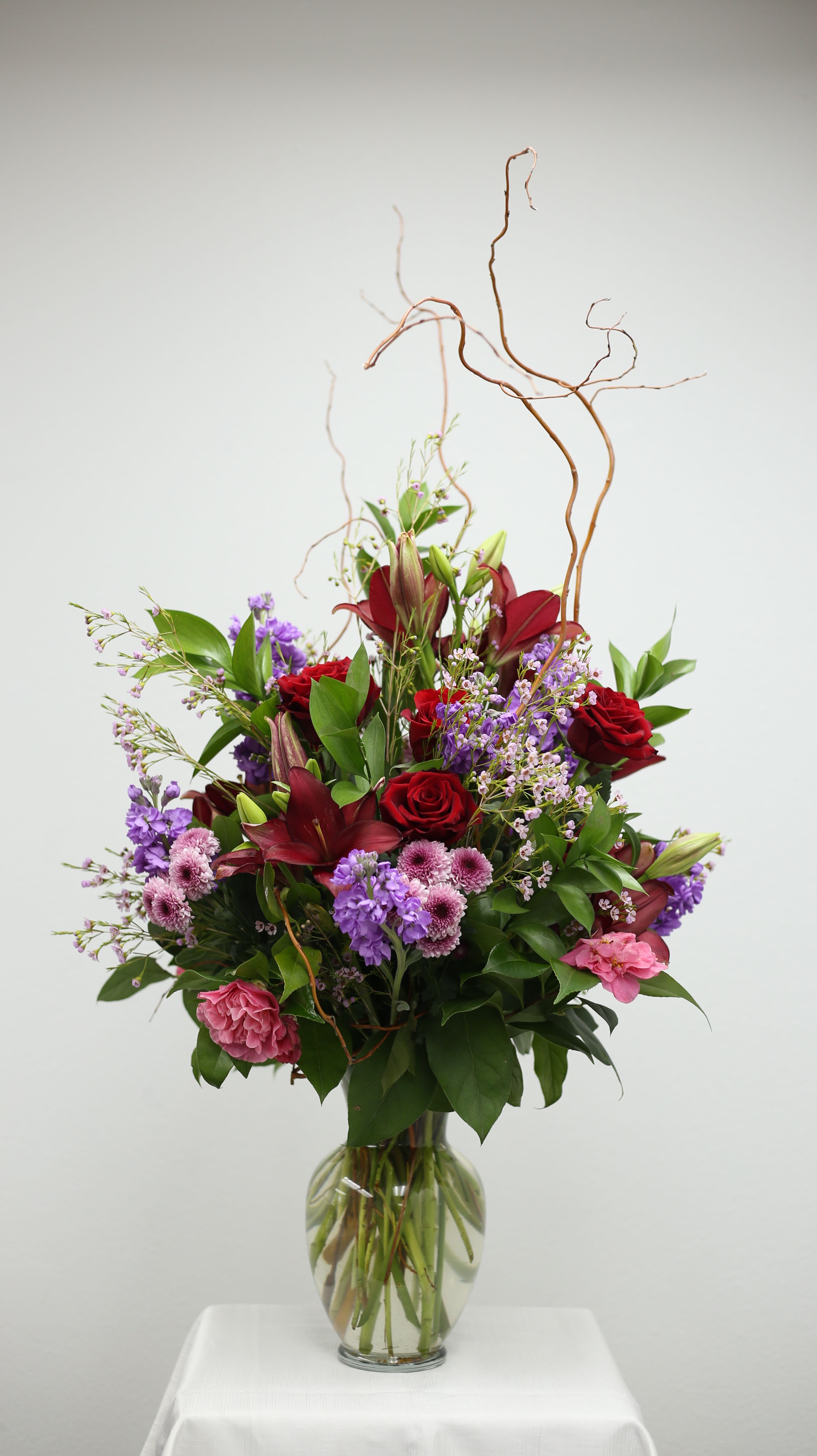 Enchanted Garden Elegance Enchanted Garden Elegance - This stunning floral arrangement captures the charm of a blooming garden with a rich blend of deep red roses and lilies, soft lavenders, cheerful pink carnations, and delicate flowers in shades of purple and white. Nestled with natural greenery and curly willow accents, this bouquet brings a sense of height, elegance, and natural beauty—perfect for special occasions or a refined touch to any space.