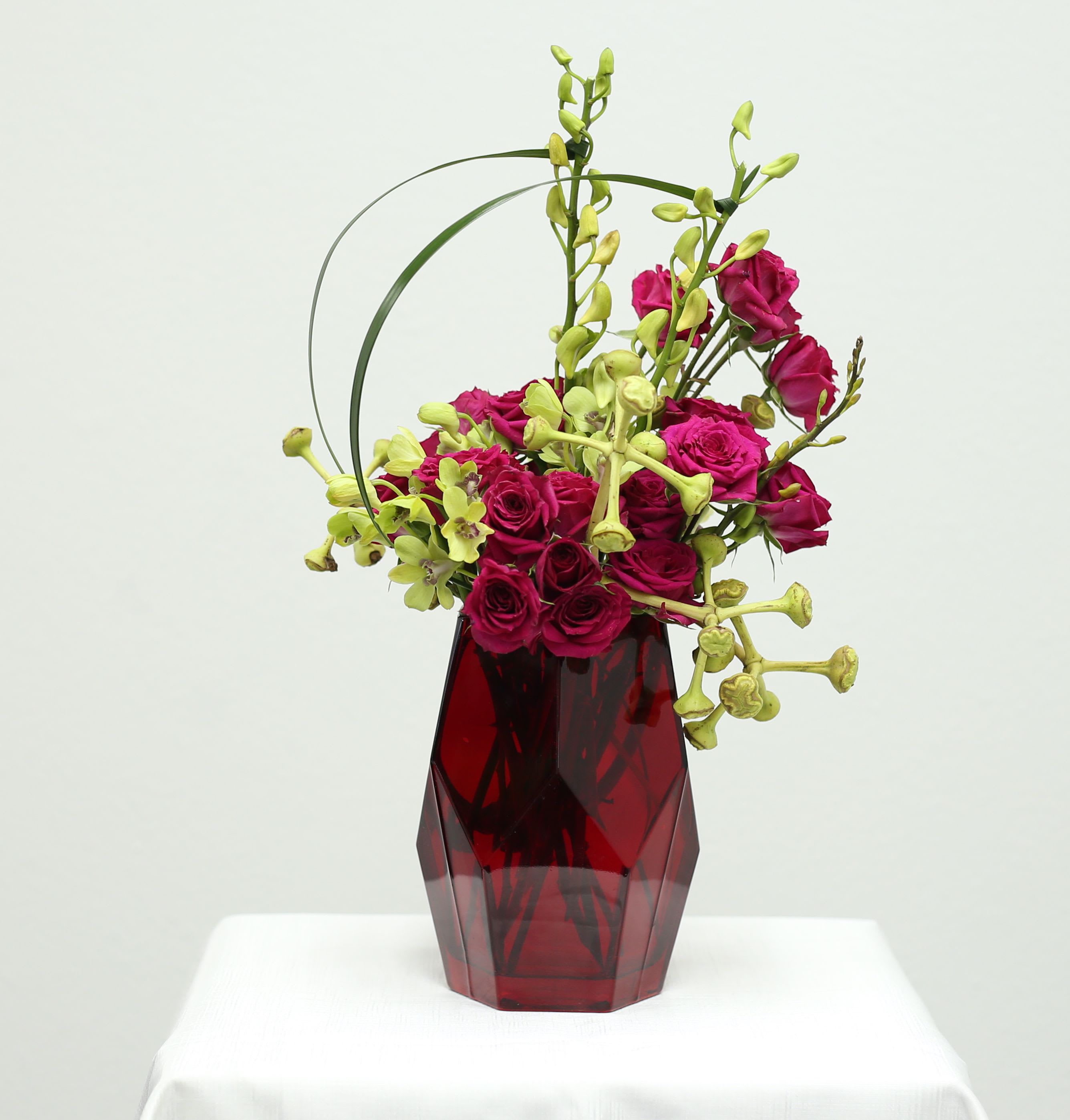 Magenta Grace Magenta Grace - Modern sophistication meets floral elegance in Maenta Grace. Deep magenta spray roses and pale green orchids cascade gracefully, accented with artistic arcs of bear grass and sculptural seed pods. This asymmetrical design strikes a perfect balance between bold color and minimalist form—ideal for upscale events, romantic gestures, or as a chic décor statement.