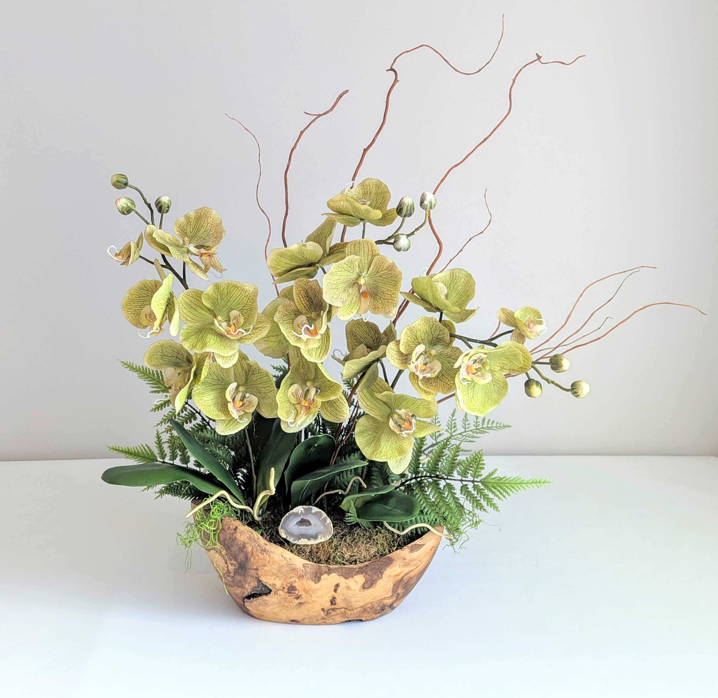 Silk Phalaenopsis Orchid Dish Garden w/Agate Geode - Green  - This custom designed &amp; one of a kind,  exquisite orchid dish garden features everlasting faux orchids set in a rich, rustic olive wood bowl. The design showcases a harmonious blend of natural textures and vibrant greenery. Lush, realistic fern greens are layered throughout, adding a soft and fresh feel. Graceful willow branches arc elegantly above, lending height and fluidity to the composition. At the heart of the garden, lush, vibrant green orchids stand out with their lifelike appearance. Crafted from high-quality artificial flowers, these orchids mimic the intricate details and vivid color of their real counterparts, ensuring long-lasting beauty that doesn’t fade over time.   Soft green moss adds an earthy element, while a natural agate geode with druzy center bring a touch of sparkle and intrigue. The geodes’ natural, crystalline formations beautifully contrast the organic greenery, enhancing the overall visual appeal. The combination of the lush green orchids, delicate ferns, flowing willow branches, and earthy moss creates a serene, botanical landscape that captures the beauty of nature in a refined, low-maintenance form. The olive wood bowl, with its smooth texture and rich grain, completes the piece, offering a warm, rustic touch that elevates the entire design. This dish garden is a perfect balance of natural elegance and enduring beauty, making it an ideal showcase piece of  decor for both home &amp; office.   Measures 27&quot; tall and 27&quot; wide Orientation: One Sided