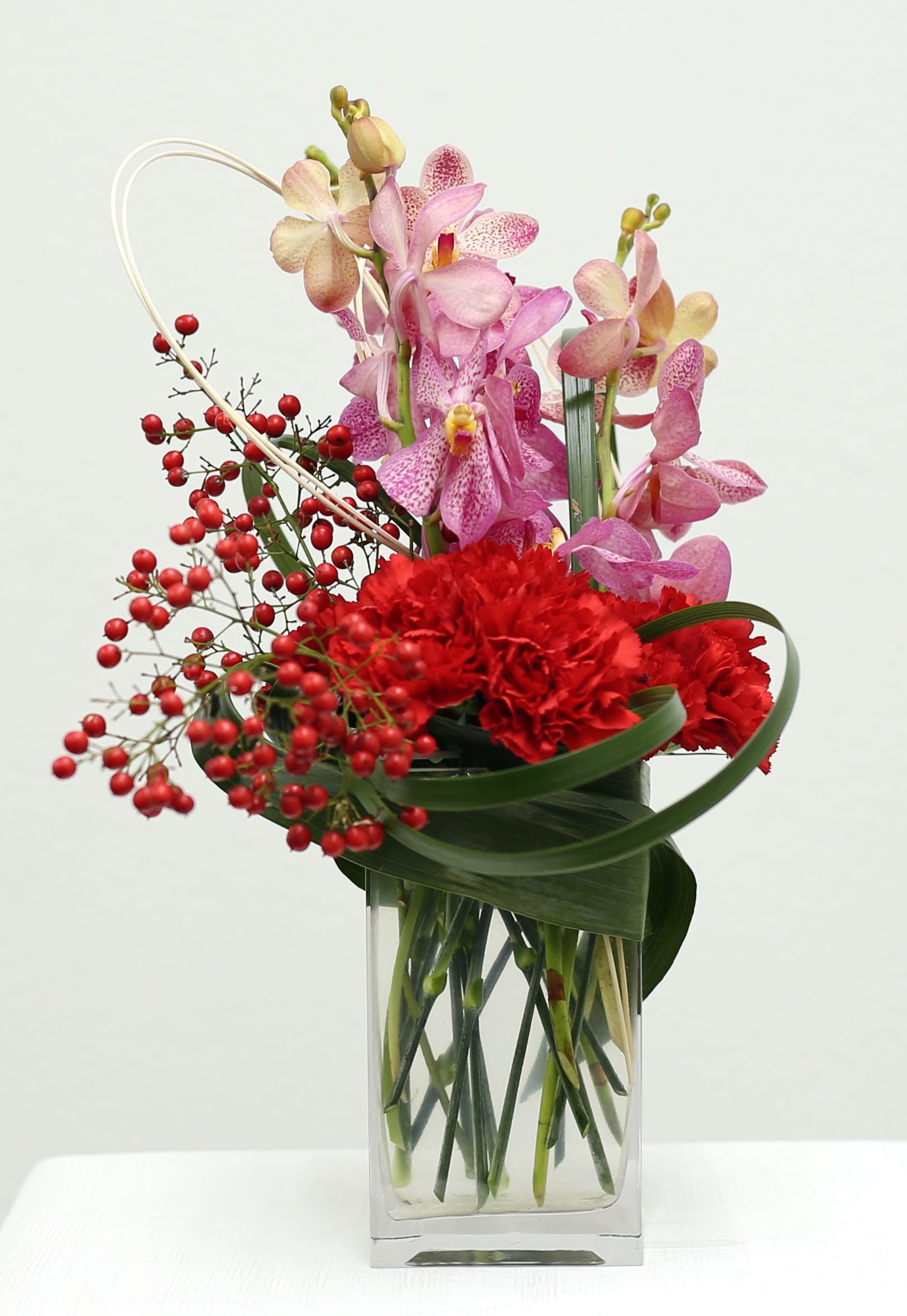 Crimson Whisper Crimson Whisper - Discover the beauty of this one-of-a-kind floral arrangement. Delicate pink orchids and vibrant red carnations are thoughtfully combined with sculptural greenery and whimsical red berries, creating a truly eye-catching display. Presented in a clear glass vase, this arrangement is a perfect gift or a stunning addition to your own space. Arrives fully assembled and ready to bring joy.