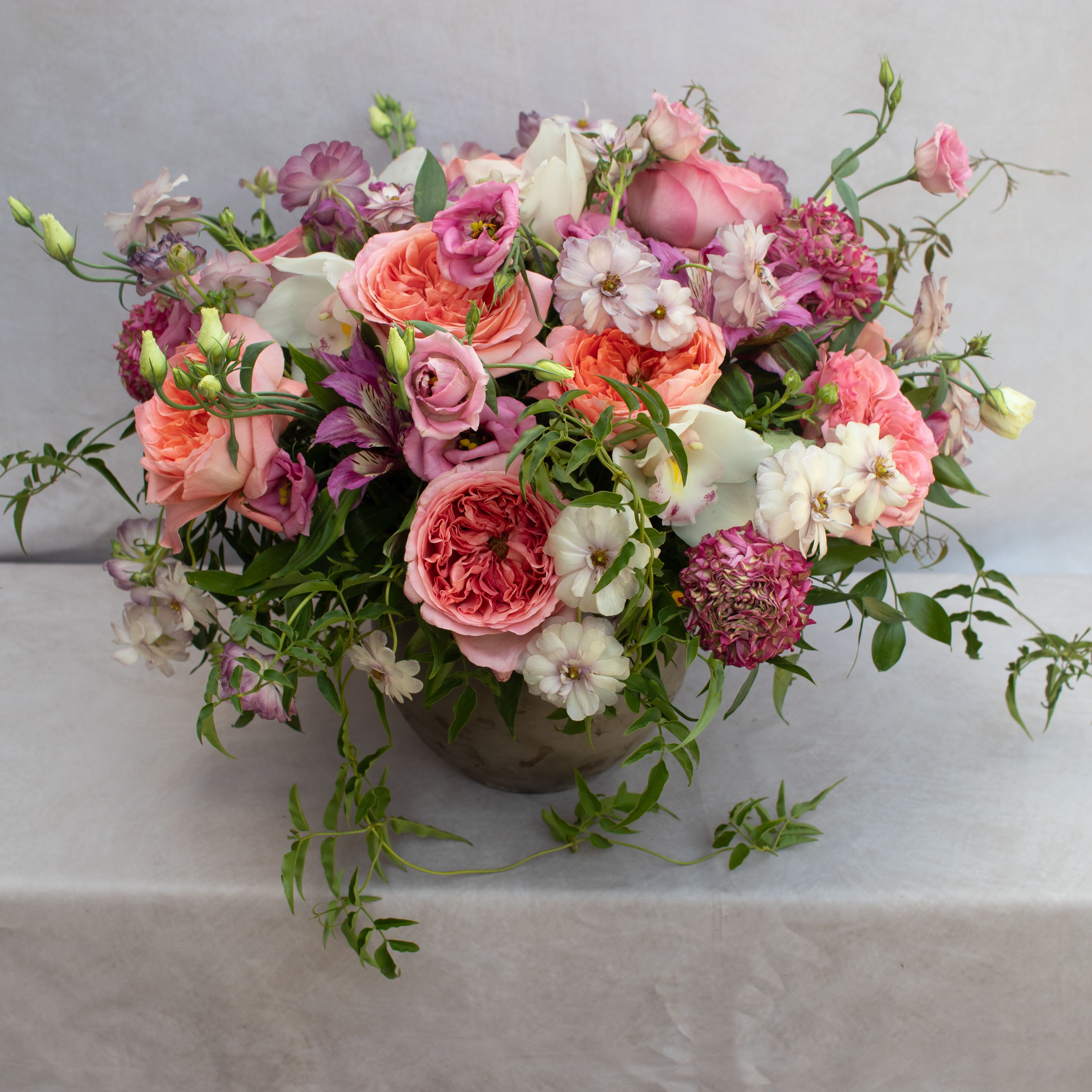 GLORIOUS GARDEN GLORIOUS GARDEN - GARDEN ROSES, DIFFERENT VARIETIES OF RANUNCULUS, LISIANTHUS, ALSTROMERIAS, TEXTURAL GREENERY, AND CYMBIDIUM ORCHID BLOOMS. ARRANGEMENT IS ABOUT 10X10 DESIGNED IN A 6X6 CONCRETE CONTAINER.