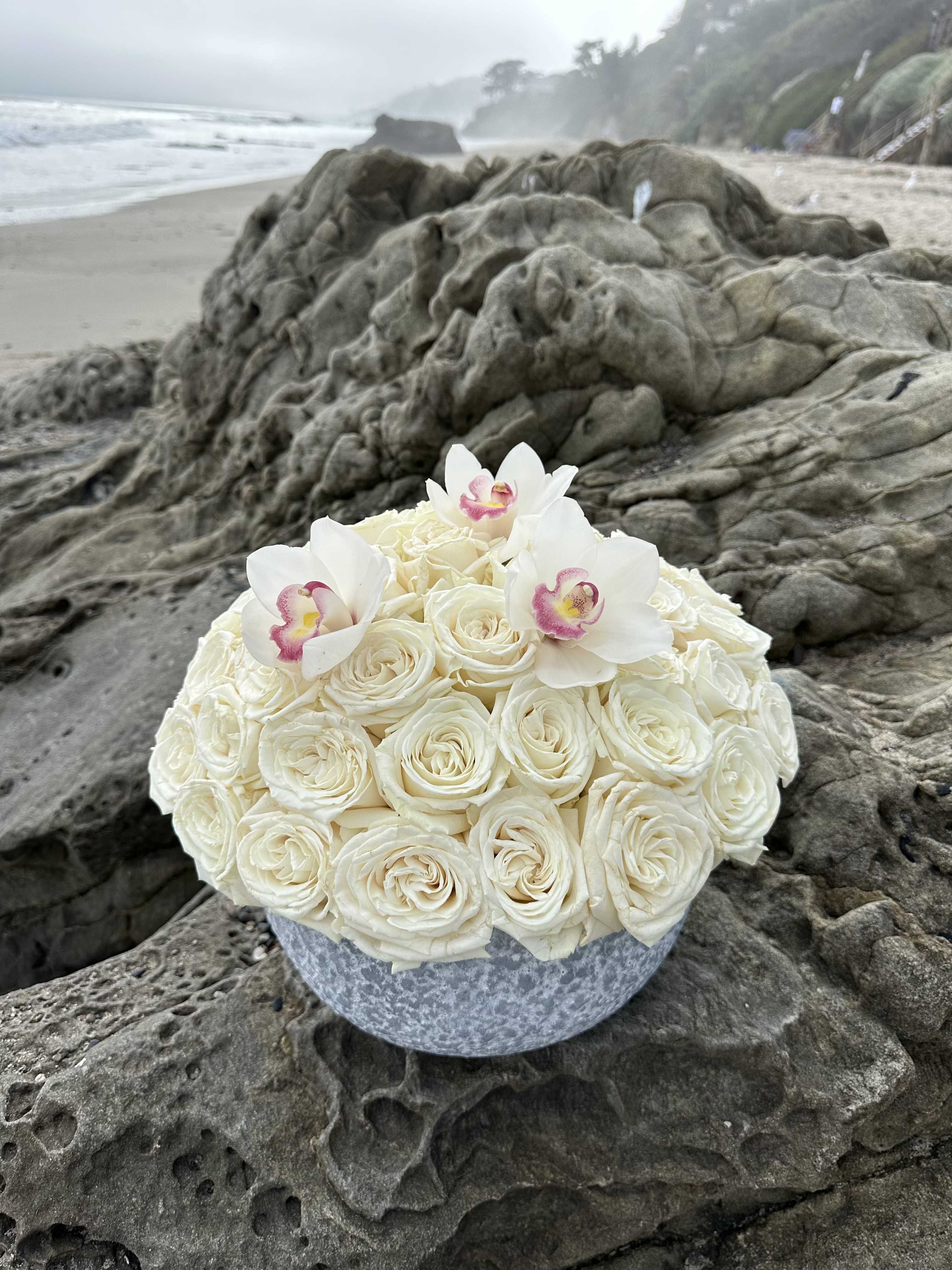 Moonlight Sonata - The purity of white roses and the refined beauty of orchids blend into a graceful composition that radiates harmony and lightness. This duo symbolizes elegance, respect, and sincere emotion.  Presented in a vase, the arrangement becomes more than just a bouquet — it’s a complete decorative accent, perfect as a gift, for a wedding, or to complement a calm and sophisticated interior. The standard size is 50 roses and 3 orchids.