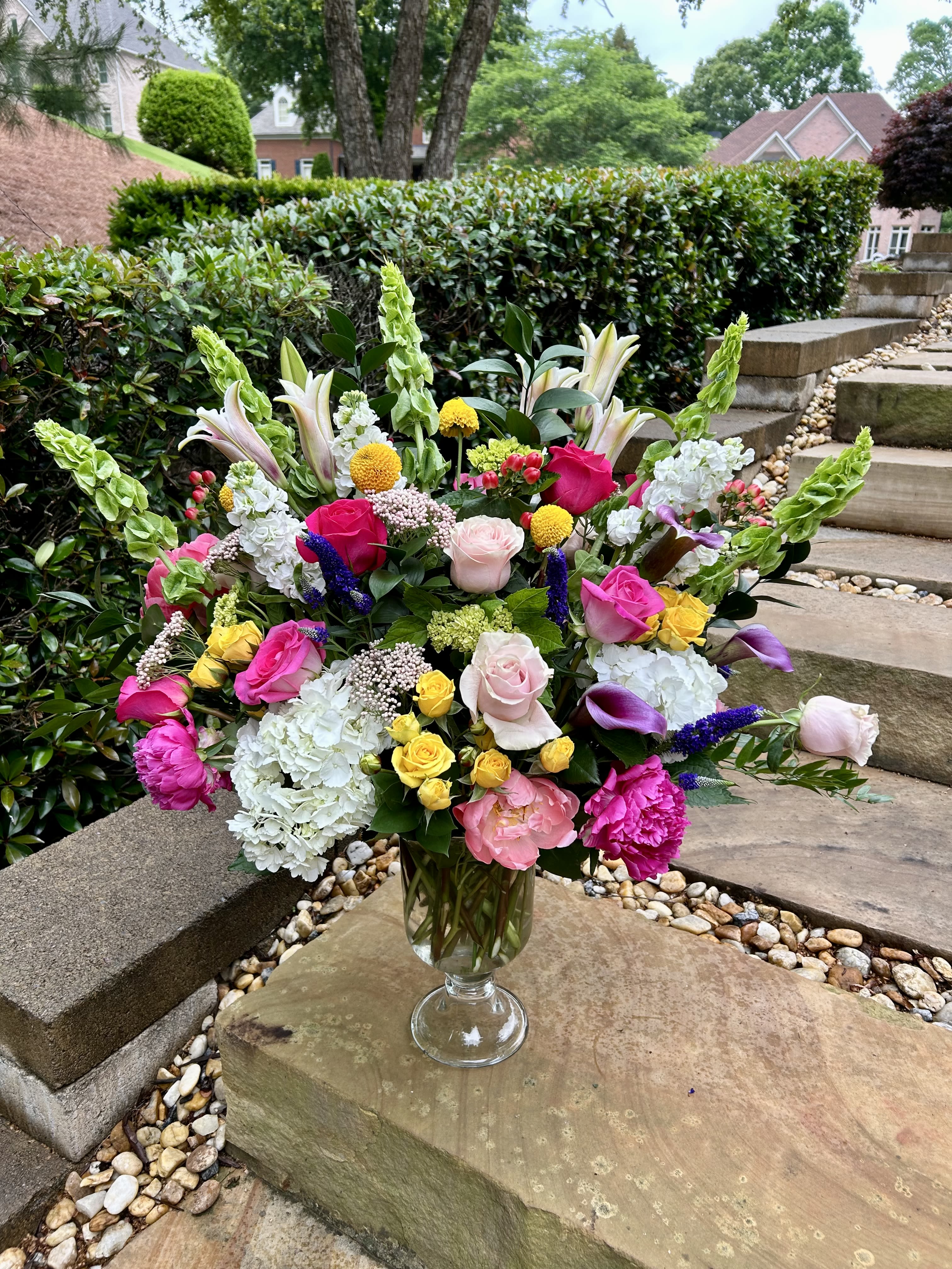 Blushing Symphony - Blushing Symphony is a stunning floral overture of color and elegance. Lush hot pink and soft pink roses and peonies take center stage, harmonizing beautifully with the cloud-like texture of white and green hydrangeas. Accents of cheerful yellow and rich purple filler flowers add a vibrant rhythm, while the tall, sculptural Bells of Ireland lend a graceful, architectural finish. This grand arrangement is a celebration of beauty and balance—perfect for making a bold yet refined statement in any space.