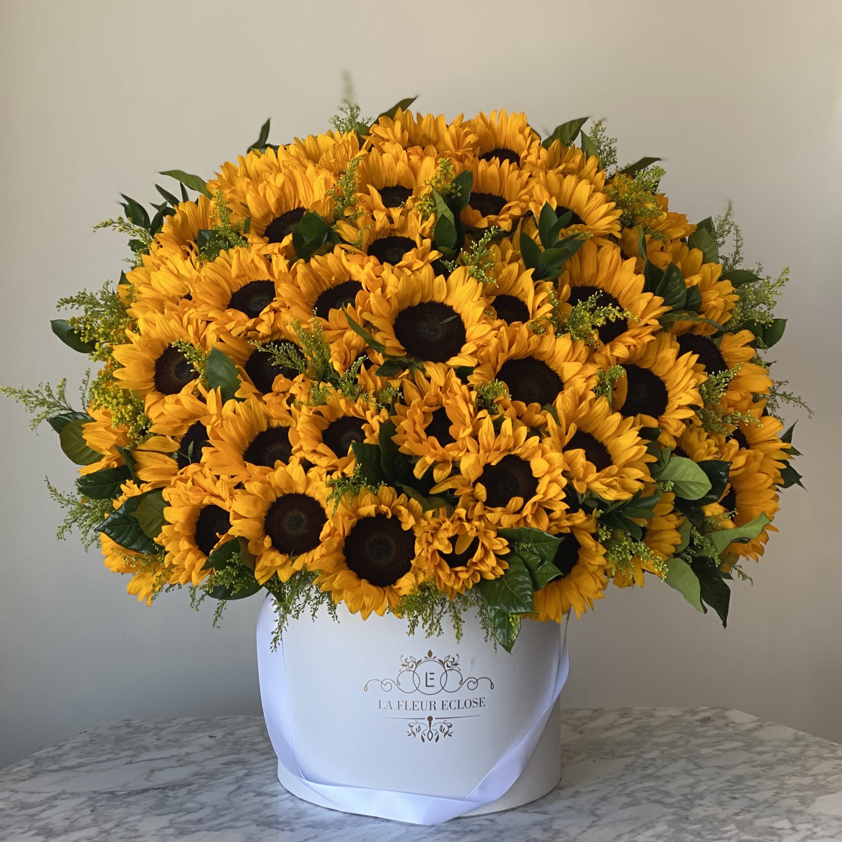 Sunflower Garden - This flower arrangement features bright and cheerful sunflowers elegantly presented in a pristine white velvet box. The vibrant yellow petals of the sunflowers contrast beautifully against the soft white velvet, creating a visually striking and luxurious display. The combination of the sunny hues of the sunflowers and the purity of the white velvet evokes a sense of joy, freshness, and elegance.  The overall aesthetic is both charming and stylish, offering a blend of natural beauty and opulent design.