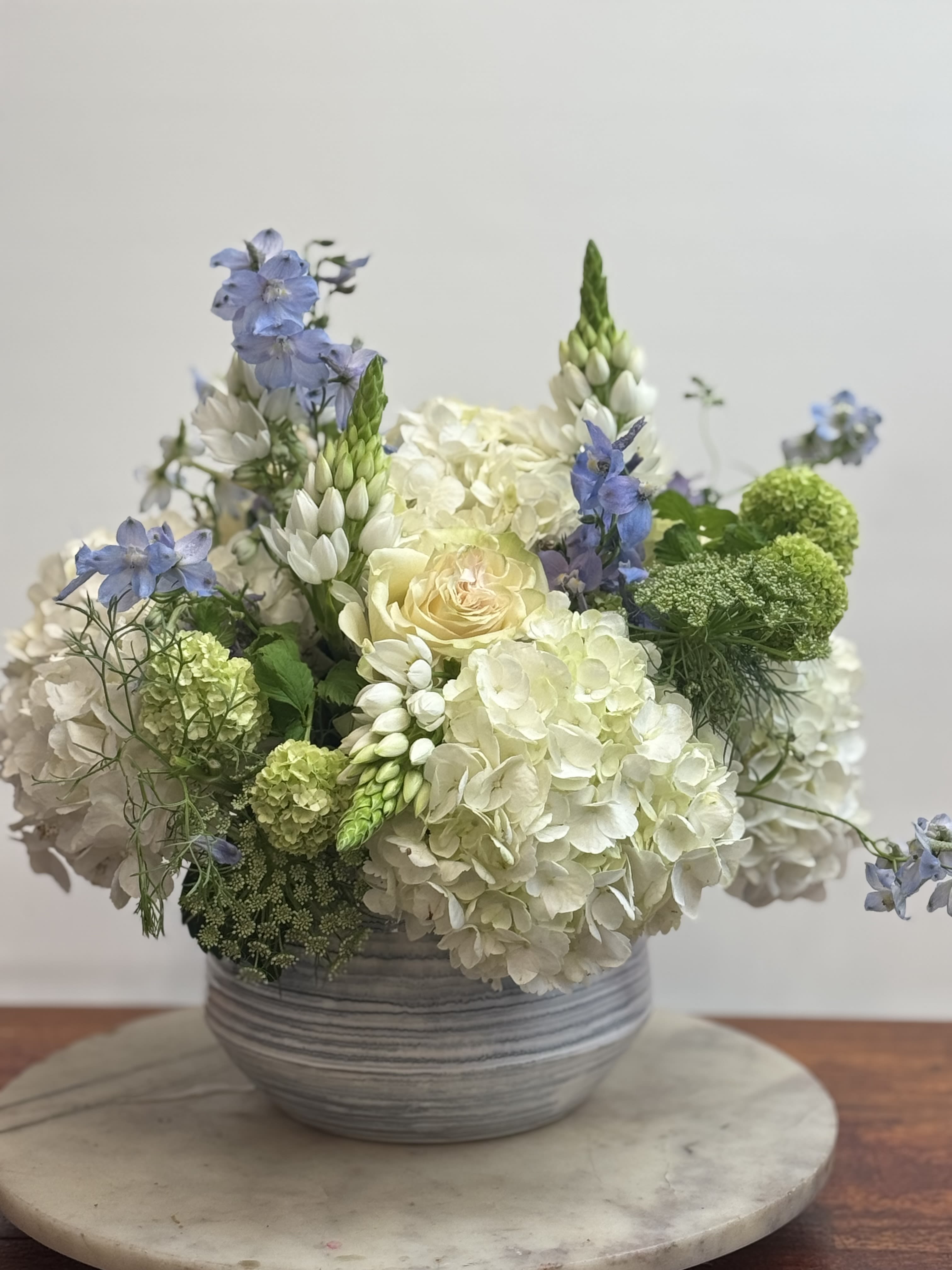 Luba’s Whisper Luba’s Whisper - Designed by our florist Jane in loving honor of her mother, this serene arrangement features soft white hydrangeas, delicate roses, lisianthus, green viburnum, and airy blue delphinium. A gentle, graceful composition that speaks to the quiet strength, beauty, and calm presence of a mother’s love.