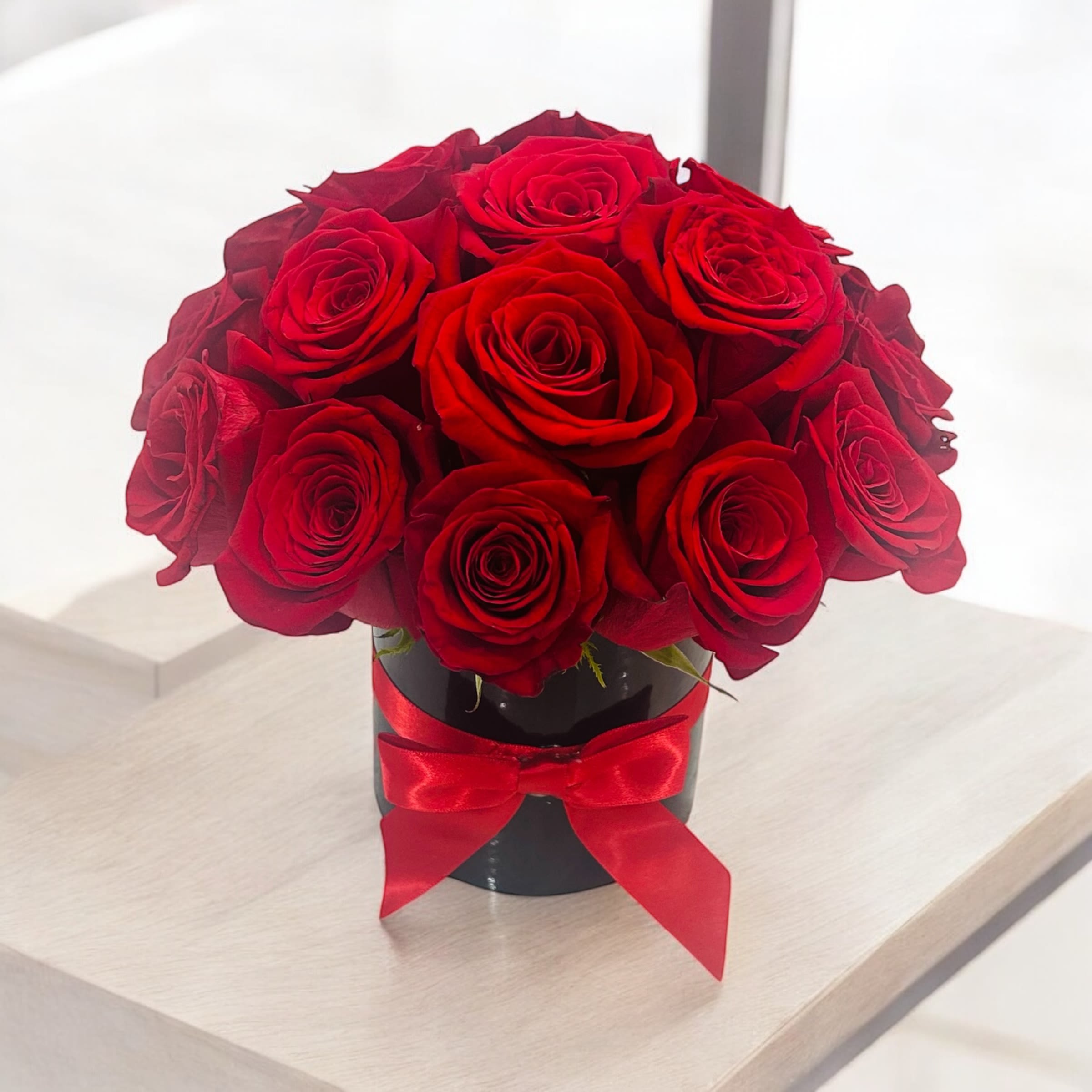 Cutie Rosie - A compact arrangement of vibrant red roses in a sleek black vase. The roses are neatly gathered, accented with minimal green foliage, creating a simple yet elegant display. The vase’s dark tone highlights the rich red of the roses, offering a refined and modern touch.