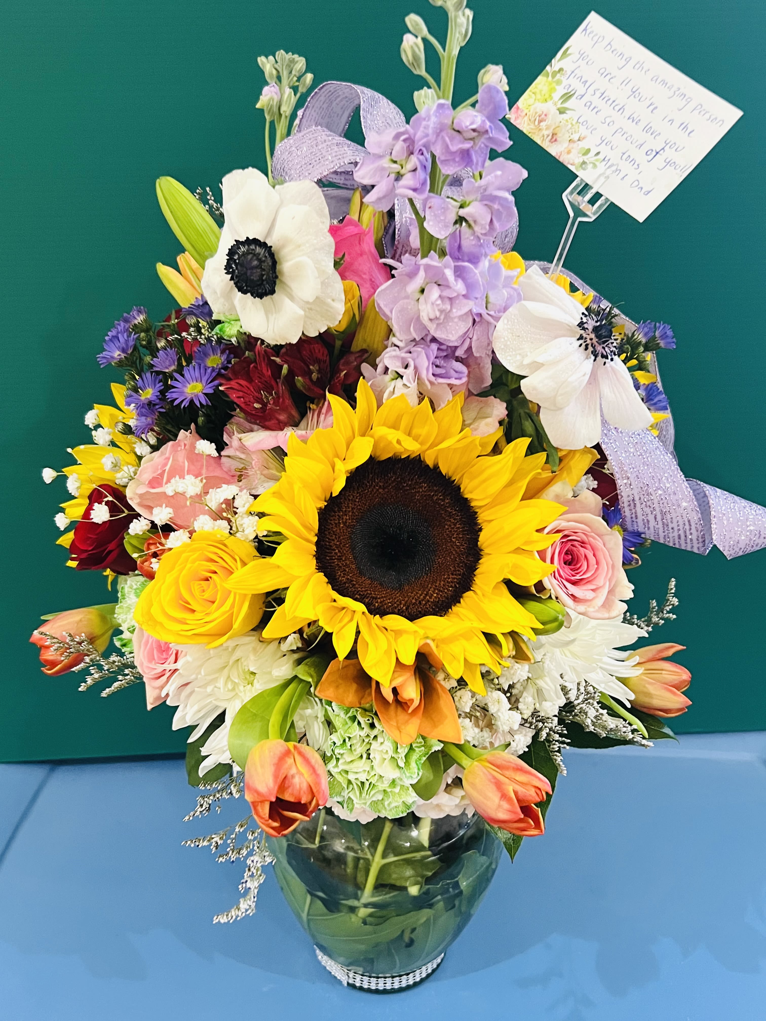 Fields Of Joy - Bursting with vibrant color and texture, this lush arrangement is a radiant celebration of nature’s finest blooms. Golden sunflowers take center stage with their bold, cheerful faces, exuding warmth and positivity. Surrounding them are velvety roses in shades of blush and coral, adding depth and romance. Fragrant stock brings height and softness, while elegant tulips lend a graceful curve and a whisper of springtime charm.