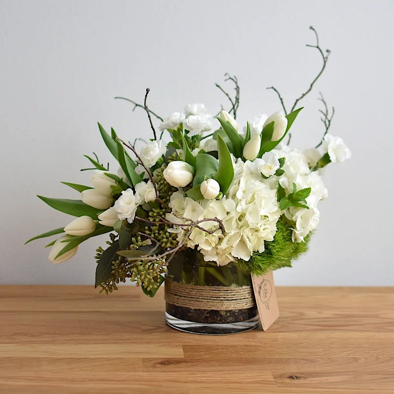 White Garden -  Are you ready to be enchanted by the beauty of our &quot;White Garden&quot; floral arrangement from Hidden Door Floral Studio? This captivating creation exudes an air of modern elegance with its asymmetrical design. It is carefully curated to captivate the senses and elevate any space with its ethereal beauty.   Immerse yourself in a harmonious blend of stunning white hydrangeas, delicate white tulips, white mini carnations, and vibrant green dianthus. Each flower contributes its unique characteristics, resulting in a mesmerizing tapestry of textures and colors that are both soothing and invigorating.  The exquisite white hydrangeas are taking center stage, their lush, billowy blooms exuding timeless beauty and purity. Symbolizing heartfelt emotions, these graceful blossoms are the perfect choice for expressing love, gratitude, or admiration in a modern and elegant way. Complementing the hydrangeas, the delicate white tulips add a touch of sophistication and grace with their slender stems and gentle petals. Evoking a sense of refinement and elegance, these blooms from Hidden Door Floral Studio elevate the arrangement to new heights.  The white mini carnations, with their ruffled petals, infuse this exclusive floral art with a touch of whimsy and playfulness. These blossoms represent fascination and admiration and contribute a modern twist to the overall composition, creating a sense of effortless charm.     
