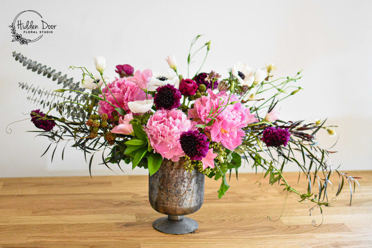 Angel Wings Angel Wings - Step into a world of ethereal beauty and grace with our stunning "Angel Wings" floral arrangement from Hidden Door Floral Studio. This captivating composition captures the delicate allure of pink peonies, the whimsical elegance of white anemones, and contrasting burgundy ranunculus and dancing lisianthus, creating a vision of heavenly enchantment. The soft and romantic pink peonies take center stage in this arrangement, unfurling their velvety petals like delicate angel wings. Each bloom exudes serenity and purity, casting a spell of tranquility wherever it is displayed. The delicate white anemones resemble celestial stars, adding a touch of ethereal charm to the arrangement. Their dainty petals and dark centers create a mesmerizing contrast, harmonizing beautifully with the pink peonies and enhancing the overall allure of the "Angel Wings" composition. Nestled in an antique-inspired glass vase, this arrangement becomes a symphony of colors and textures. The timeless elegance of the pink peonies and white anemones is further accentuated by lush seasonal greens, which cascade like wings on the sides, adding an exquisite touch to the display.