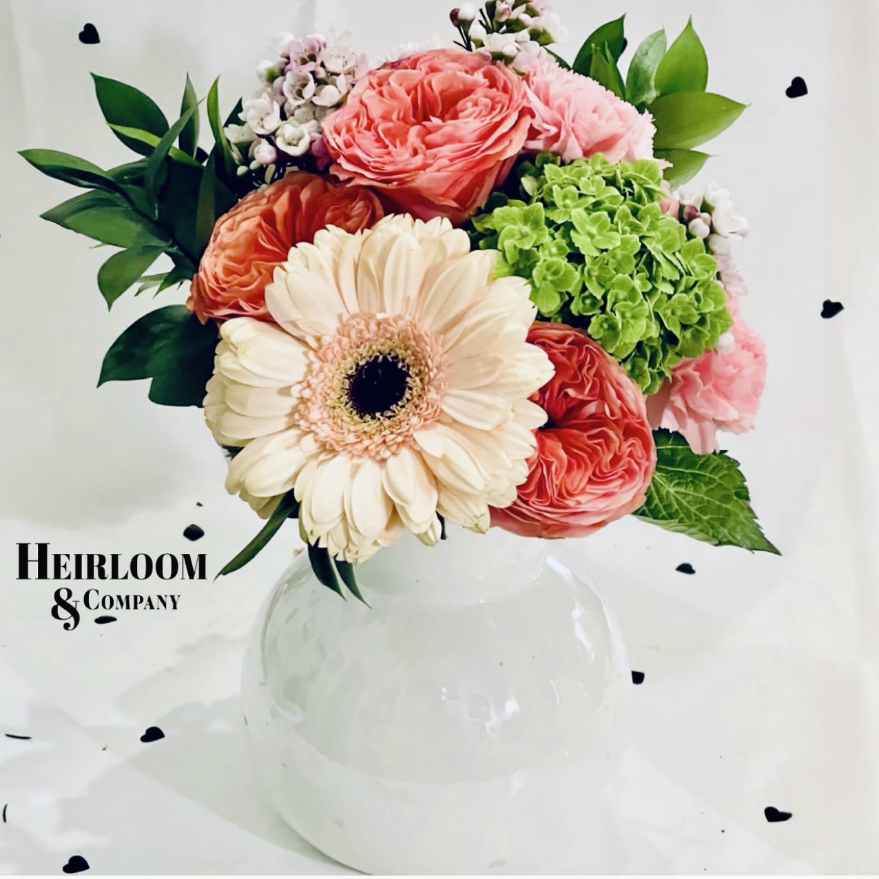 Lovebug Lovebug - This floral arrangement exudes charm and elegance, featuring a delightful mix of peachy garden roses, delicate pink carnations, fresh green hydrangeas, and a stunning cream gerbera daisy at the center. Accented with lush greenery and soft wax flowers, the bouquet is arranged in a classic white vase, making it perfect for brightening any space with its romantic and fresh aesthetic. The soft color palette and intricate textures create a timeless and graceful composition.