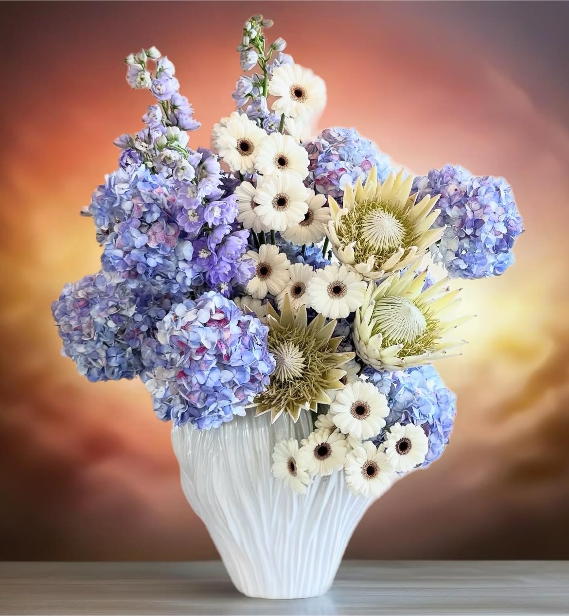 Breathing beauties  - This extraordinary floral arrangement is a stunning composition that combines vibrant Blue Holland hydrangea with striking giant white gerbera daisies. The serene blue of the hydrangeas contrasts beautifully with the crisp elegance of the daisies, creating a cheerful and sophisticated look.   At the center, the royal protea adds drama with its unique spiky texture and rich pink hues, while delicate blue delphiniums cascade gracefully, adding height and movement. Together, these exquisite blooms form a harmonious blend of colors and textures, transforming any space into a captivating sanctuary of beauty.