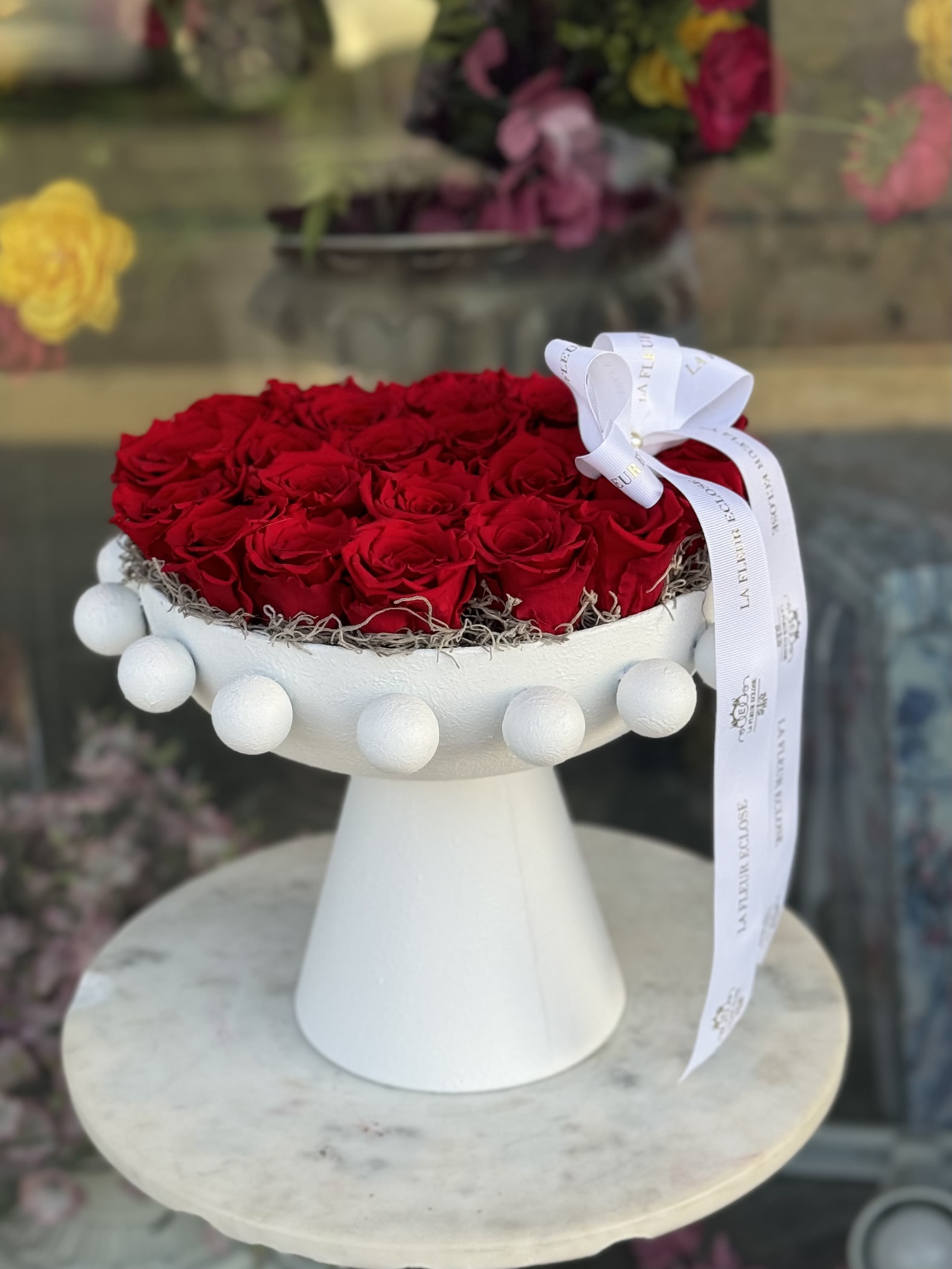 Eternal Roses on Pedestal Eternal Roses on Pedestal - An elegant crown of preserved red roses, arranged in a heart shape and set atop a sculptural white pedestal with pearl-like accents. These roses are real, carefully preserved to last up to two years — no watering or maintenance required. Perfect for a timeless love, a luxurious gesture, or an unforgettable statement of affection.