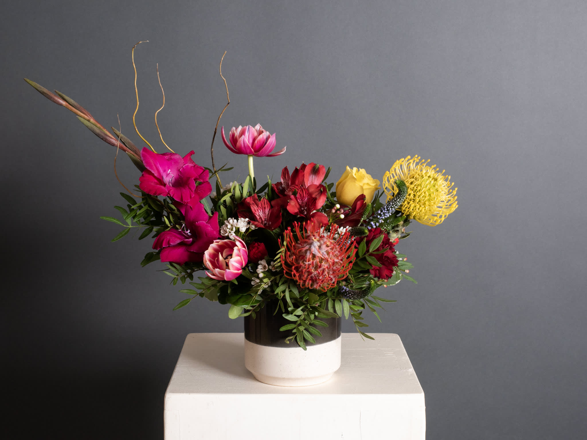 Capetown, Wanderlust Series - Ceramic Pot Arrangement - This arrangement showcases the rich, warm colors inspired by the breathtaking beauty of the iconic Table Mountain. Lush greens evoke the vibrant ecosystem that thrives above the cerulean blue of the raging ocean. The sunrise peeking over this majestic natural rock formation is reflected in the radiant orange pin cushions. The wild pink hues of gladiolus and tulip blossoms mirror the striking king protea blooms that grace the Peninsula Sandstone Fynbos. A heartfelt homage to the splendor of this unforgettable place — dreaming of the day we can return to its magic !  Product shown at $134.95 Pot size : 4.5in X 4.5in