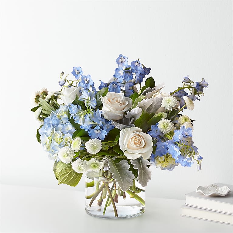 Blue Skies Bouquet - Soothing and elegant, this bouquet captures the freshness of a clear blue sky. Featuring lush blue hydrangeas, airy blue delphinium, classic white roses, and delicate white button poms, it's finished with soft greenery for a light, graceful touch. Perfect for celebrations, comforting moments, or simply bringing a little peace into someone's day.