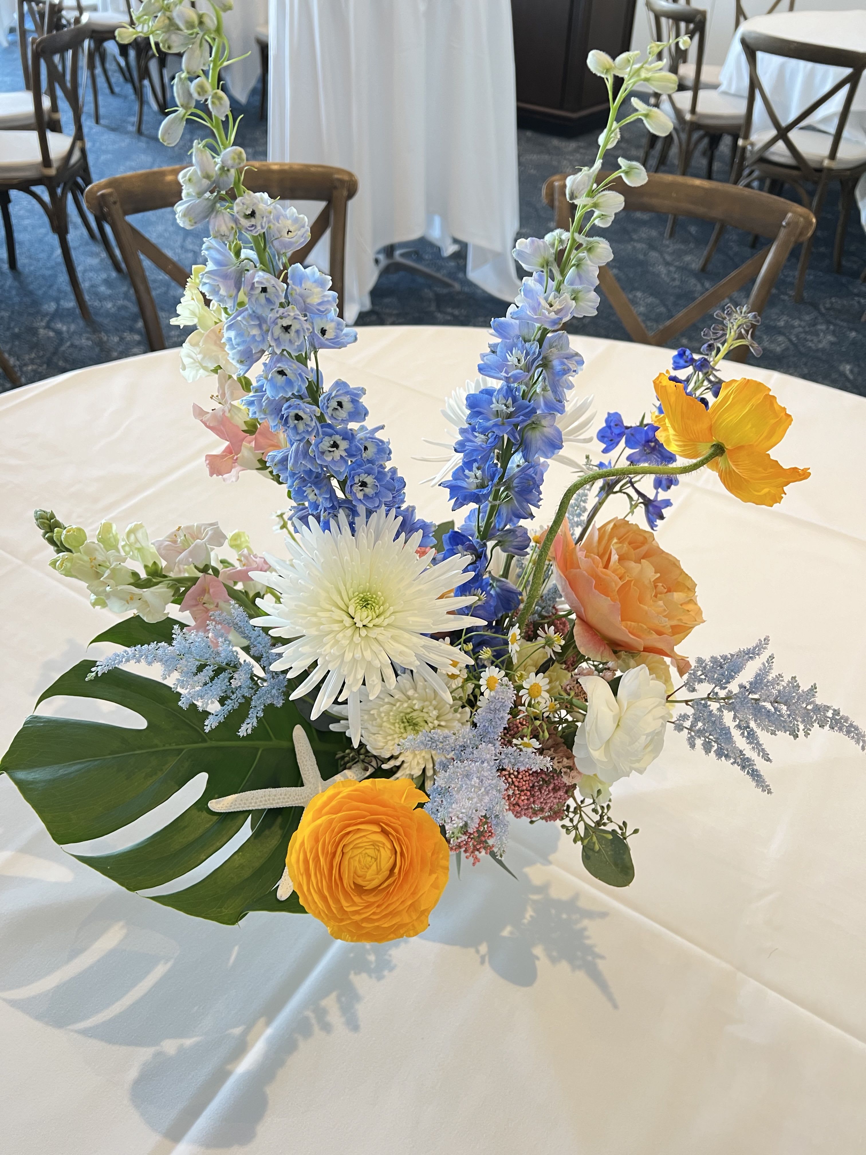 Coastal Celebration Event Arrangement - Bring the spirit of the shore to your special event with our &quot;Coastal Celebration&quot; arrangement — a bold and elegant centerpiece that captures the vibrancy of the tropics and the serenity of the sea. Featuring a lively mix of sun-kissed orange and yellow blooms, crisp white florals, and calming ocean blues, this design is accented with lush tropical foliage and a charming sea star detail for a finishing touch of coastal magic.  Whether you're hosting a beachside wedding, a summer soirée, or a nautical-themed celebration, this arrangement sets the tone with natural elegance and island-inspired flair. Each piece is handcrafted to reflect your unique event, evoking a feeling of warm breezes, salty air, and sunlit waves.  Custom sizes and variations available for large-scale installations or table centerpieces.  Available for delivery, pick up or special events in Newport Beach area.  Specific flower variety is subject to availability.  Flower Delivery in Newport Beach, Costa Mesa, Irvine, Huntington Beach, Laguna Beach, Newport Coast, Balboa Island. 