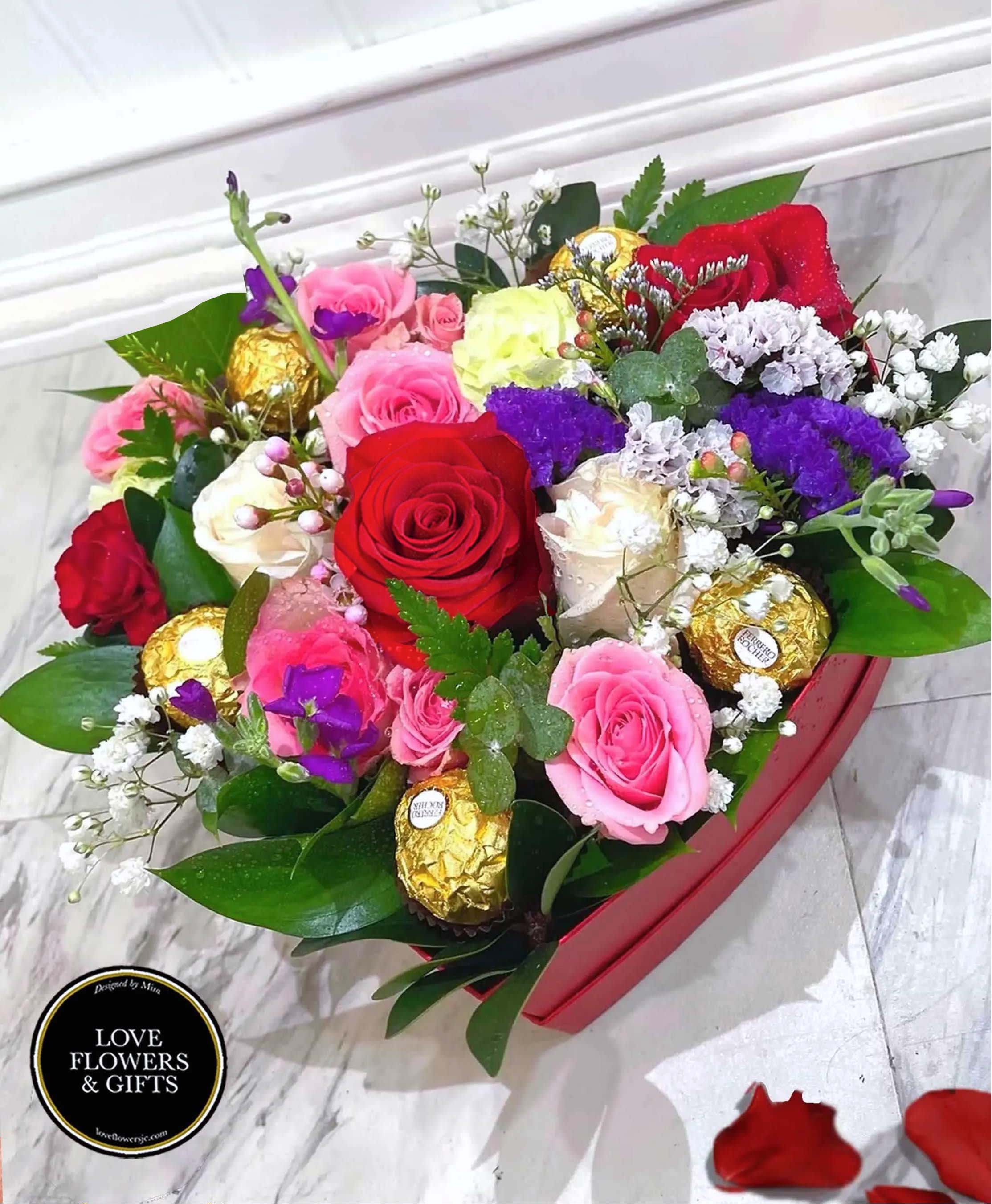 Heart-shaped box of red, pink, and white roses with purple accents and gold-wrapped chocolates.