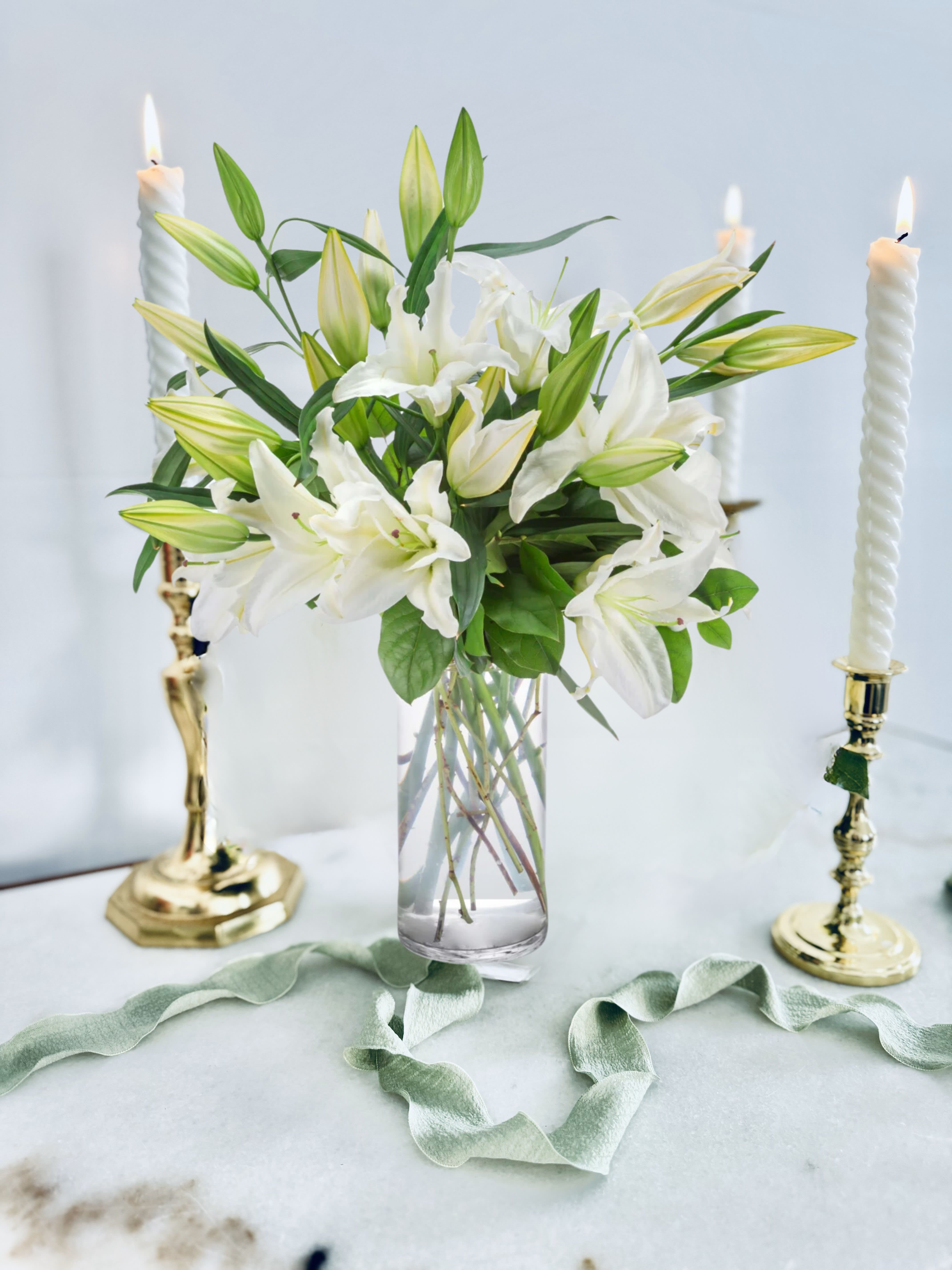 Peaceful Evening - White lilies stand tall and beautiful. Send an impactful gift to somebody whether it be for saying I am sorry or celebrating the new year. This arrangement instantly elevates any room and fills the space with that distinct sweet-smelling lily perfume.  1st Image: Standard 2nd Image: Premium  CANDLES NOT INCLUDED  As flowers are a natural product, it may be necessary to make substitutions of floral varieties on occasion due to various factors. The substituted florals will be selected based on their value, color, and overall aesthetic to match the original arrangement shown in the photos.  To maximize the longevity and aesthetic appeal of our flowers, we recommend refreshing the water in your arrangement daily. Additionally, it's advisable to keep your arrangement away from all heat sources, such as warm air drafts, direct sunlight from windows, and stoves, among others.