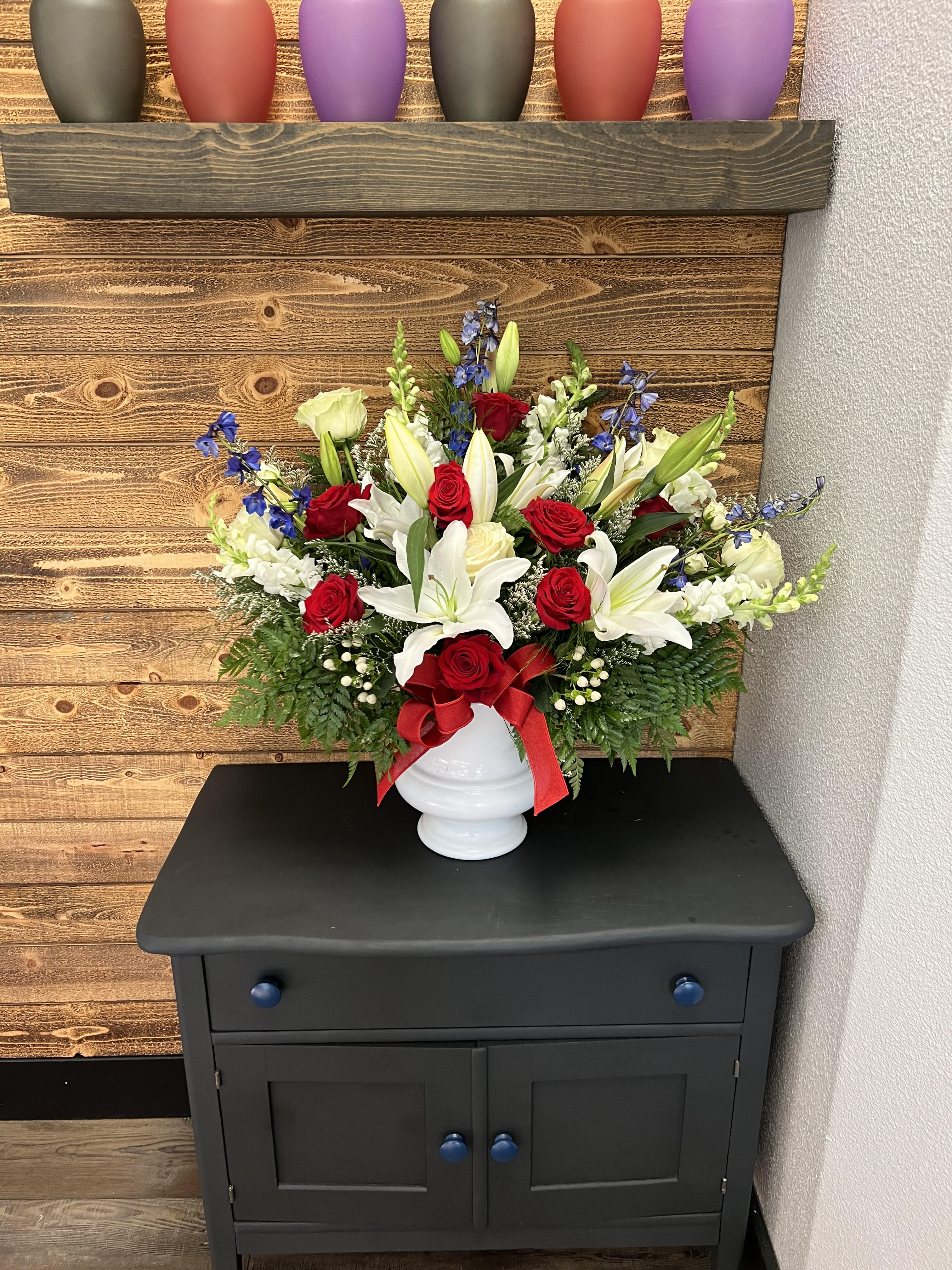With Distinction by Anderson Florist With Distinction by Anderson Florist - A mix of red, white and blue flowers such as Asiatic lilies, roses, carnations, snapdragons, chrysanthemums and blue delphinium arranged in a white urn.