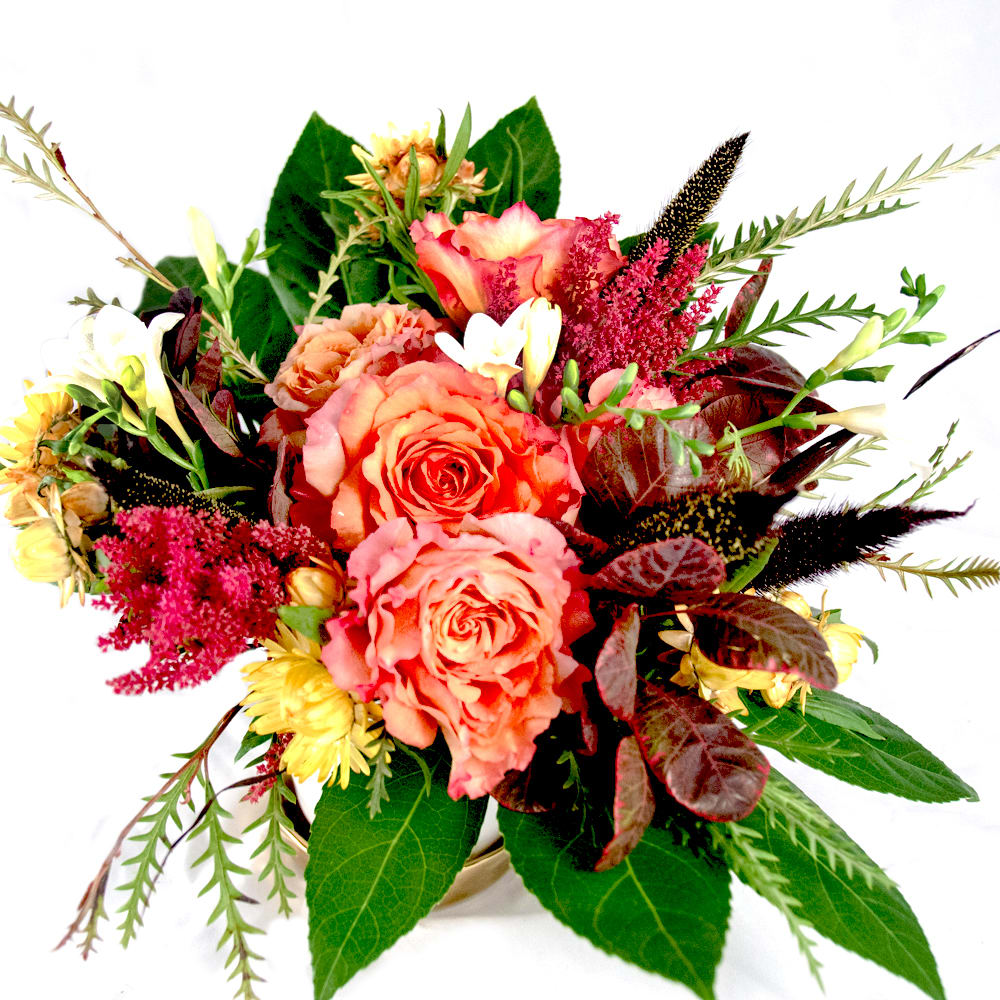 Aurora - A bombastic blossoming of colors and fragrances. Low compact design comprised of Free Spirit roses, freesia, grevillea, smoke plant leaves, bright yellows and a large size green leaf accent. (Approx. size 12&quot;w x 10&quot;h x 10&quot;d)